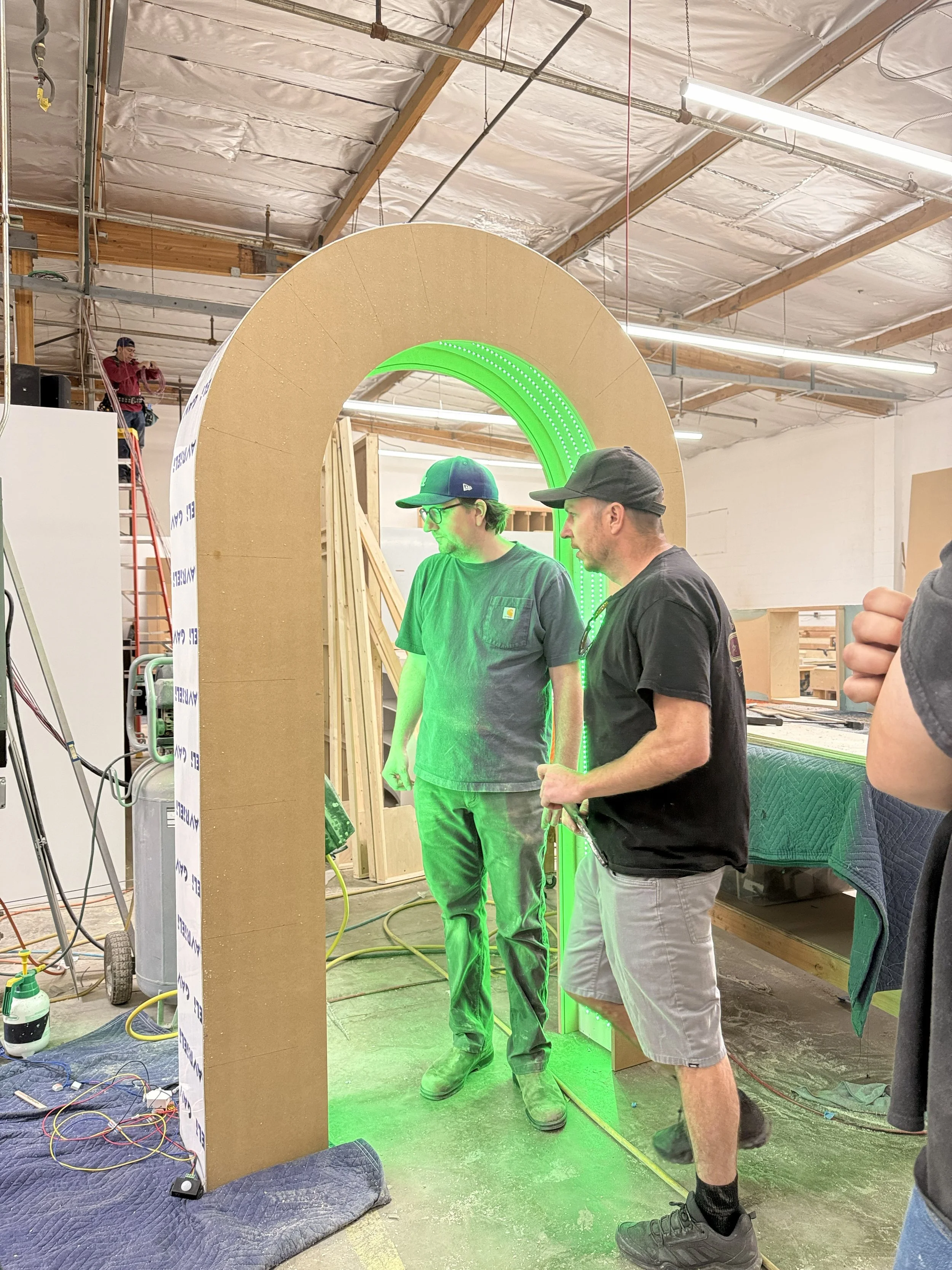 Two men standing and examining a structure with an arched opening, inside a warehouse or workshop. The structure has a green LED light detail. Tools, wires, and construction materials are visible around them.