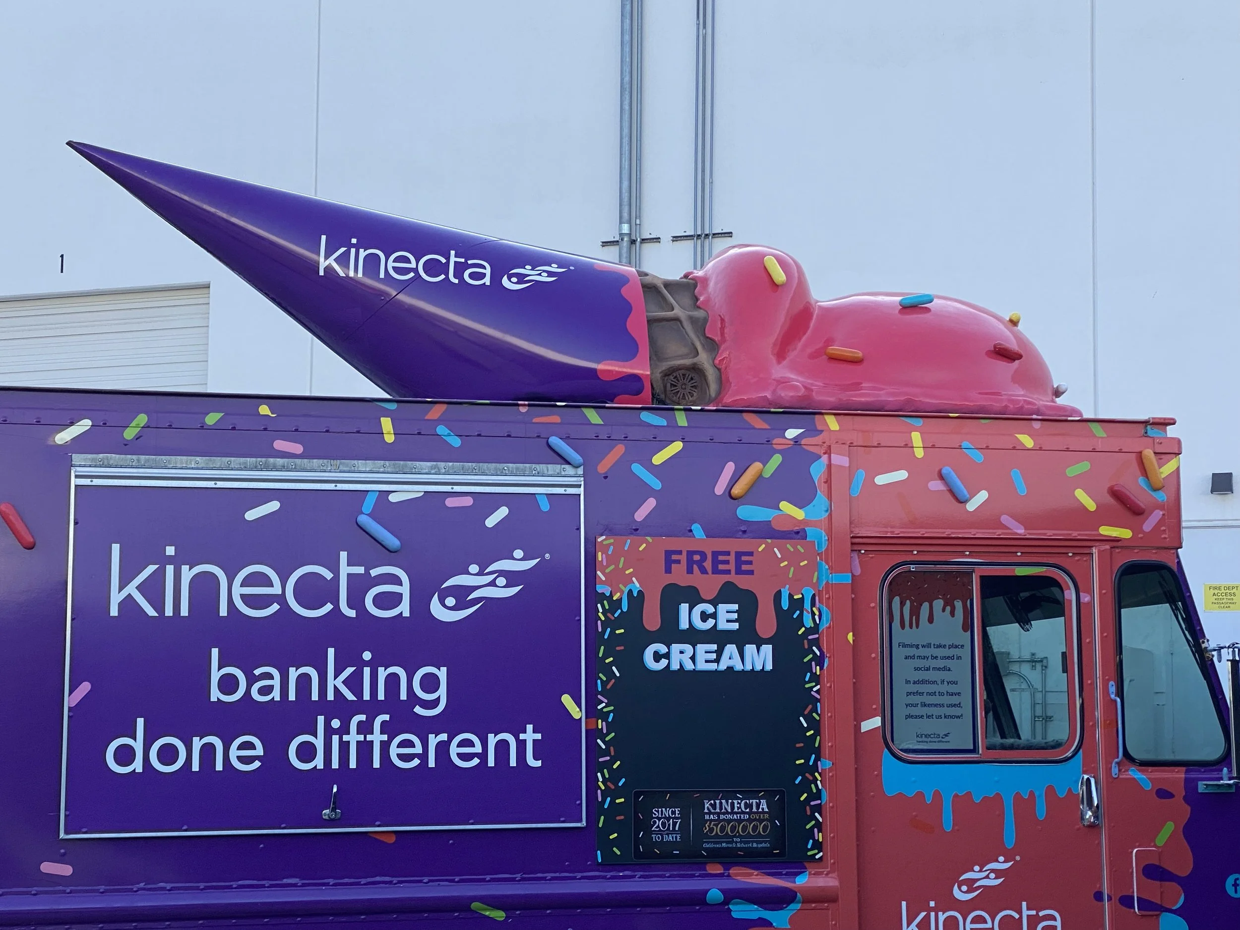 A colorful food truck with purple, pink, and multicolored sprinkles design, advertising free ice cream. The top of the truck has a large ice cream cone with pink icing and yellow, blue, and red sprinkles, with the cone displaying the 'kinecta' logo.