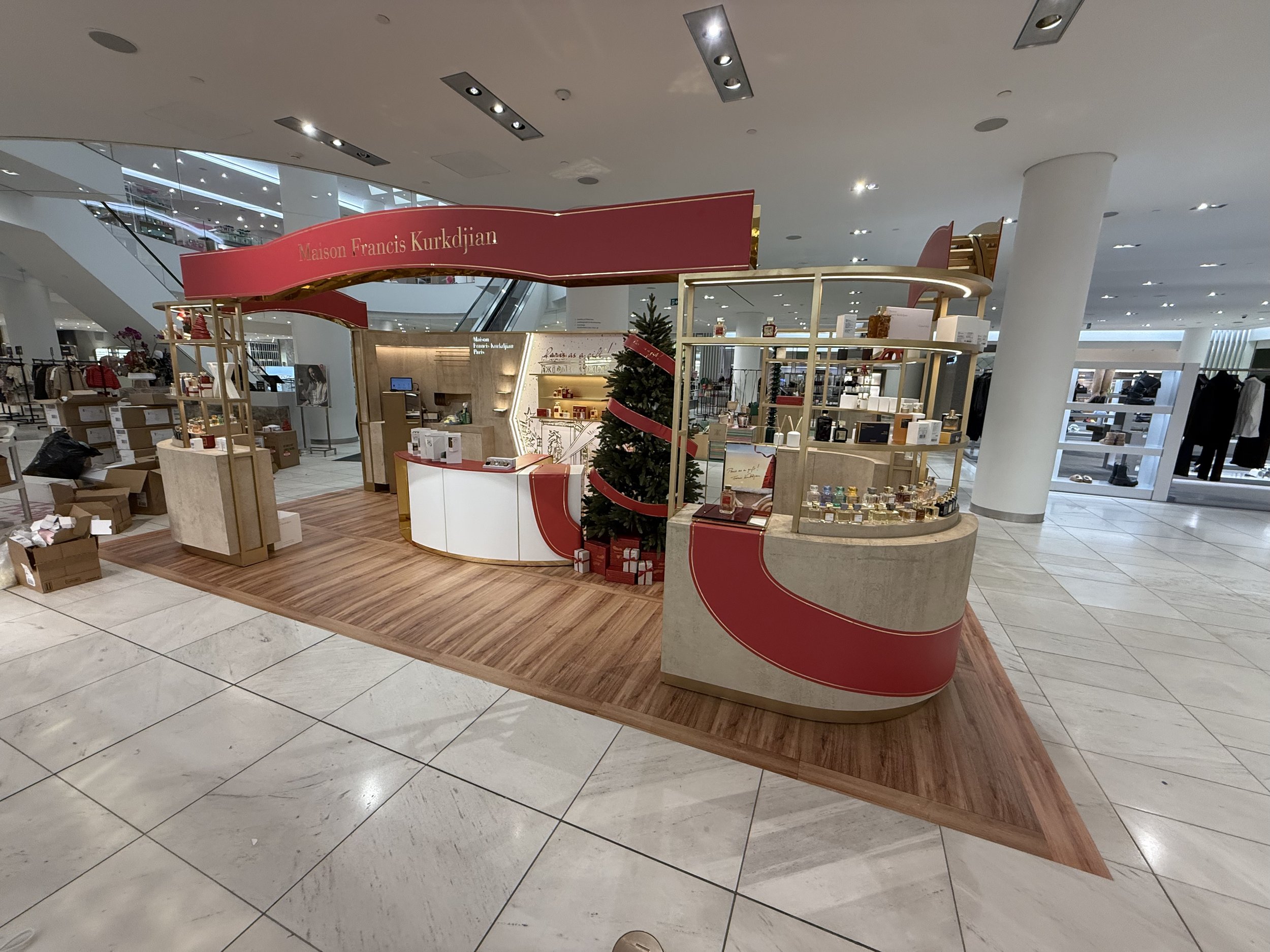 A retail display booth with a pink and red sign reading 'Maison Francis Kurkdjian' in a shopping mall, surrounded by products, a small Christmas tree, and cardboard boxes on the floor.