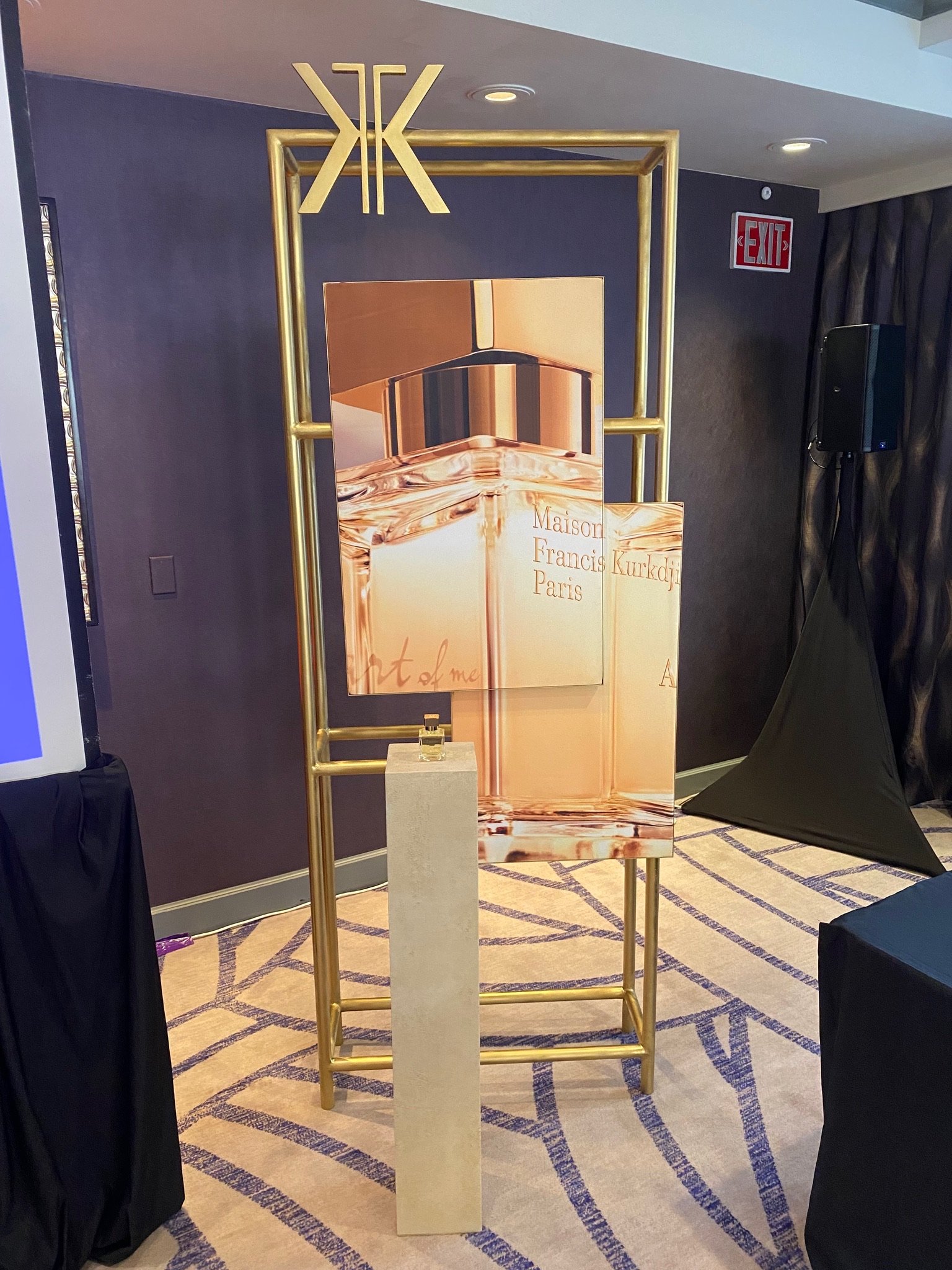 An art display with a gold metallic frame, a marble pedestal with a small object, and a promotional poster for Maison Francis Kurkdjian Paris. The setting appears to be indoors with carpeted floor and ceiling lights.