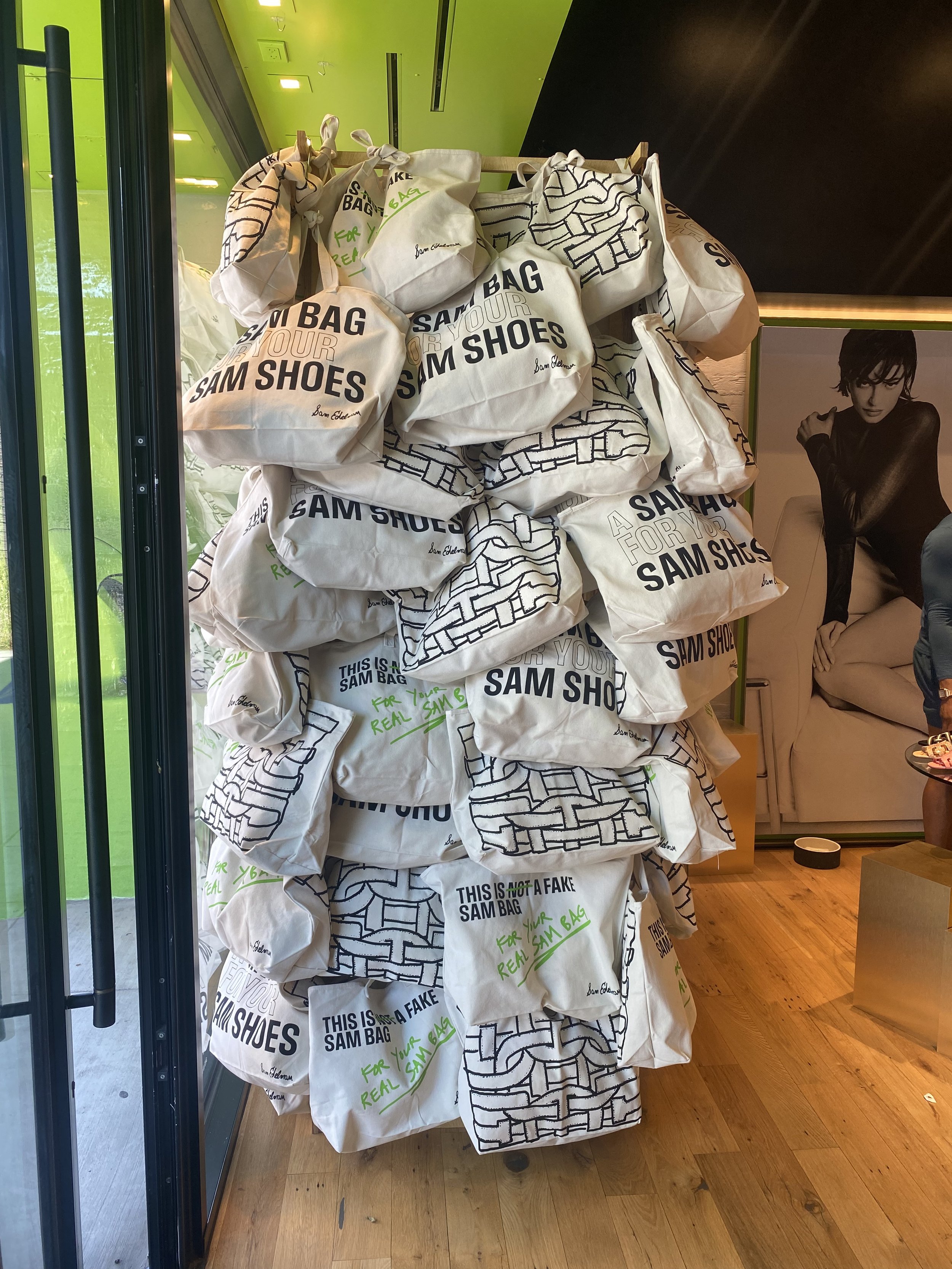 Stacked white canvas tote bags with black text reading 'This is not a fake Sam Bag' and handwritten green text, on a wooden floor in a store corner, with a black photo of a person on a wall in the background.