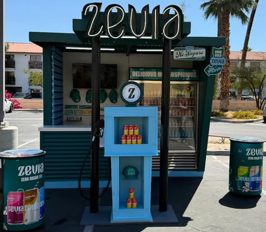 A small Zevia soda stand with a sign that says "No Sugar, Delicious, Nutritious," featuring Zevia cans on display, located in a parking lot with palm trees in the background.