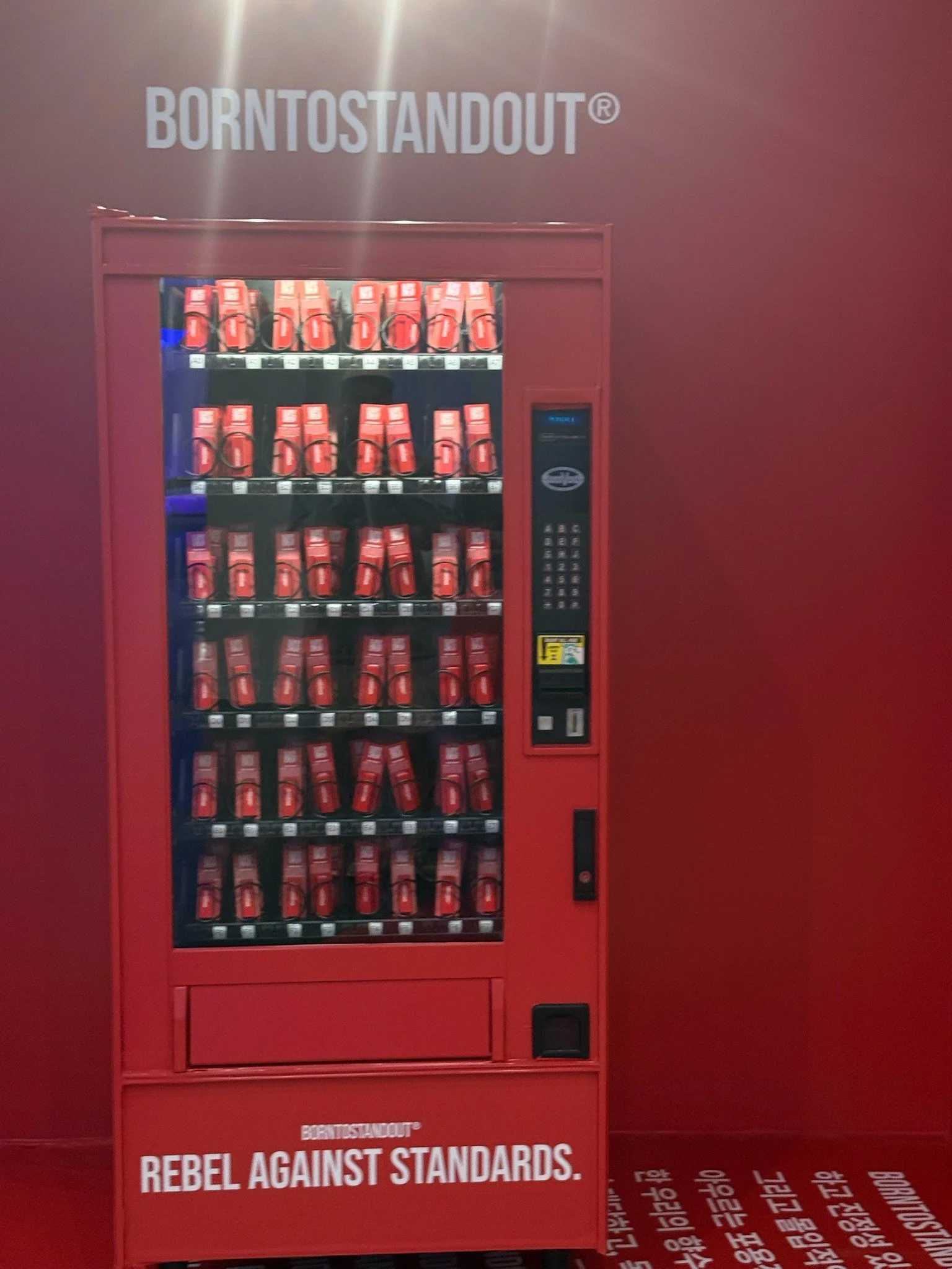 Red vending machine with multiple rows of snacks or drinks, located against a red wall with the text 'BORN TO STAND OUT' and 'REBEL AGAINST STANDARDS' printed on the red surface.
