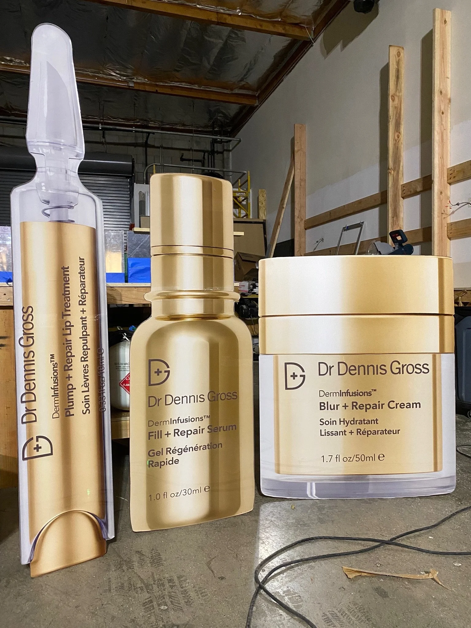 Display of various skincare products from Dr. Dennis Gross, including formulations for skin repair, firming, and smoothing, arranged against a workshop or storage space with visible wooden framing and an electrical cord on a concrete floor.