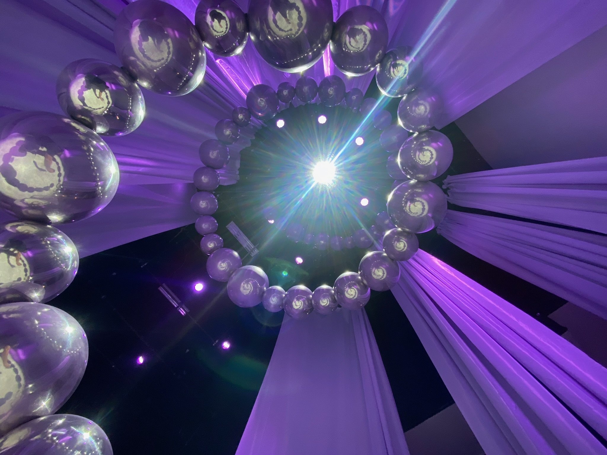 A spiral arrangement of transparent balloons with a purple reflective surface, illuminated by purple and blue lights, with a bright white light at the center creating lens flare effects.