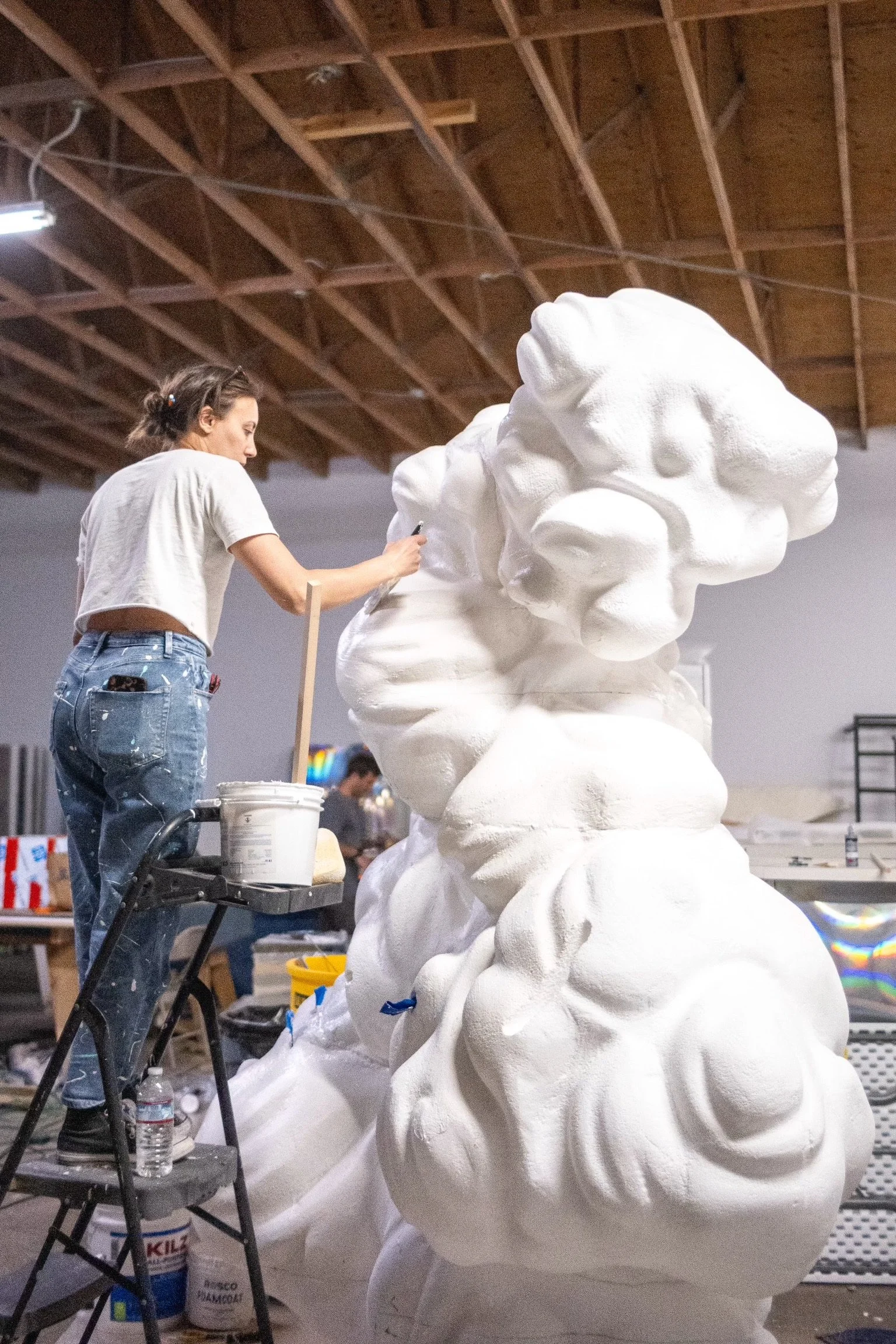 A woman working on a large abstract white sculpture in an indoor studio or workshop. She is wearing a beige t-shirt and blue jeans, standing next to a small tools and materials cart.