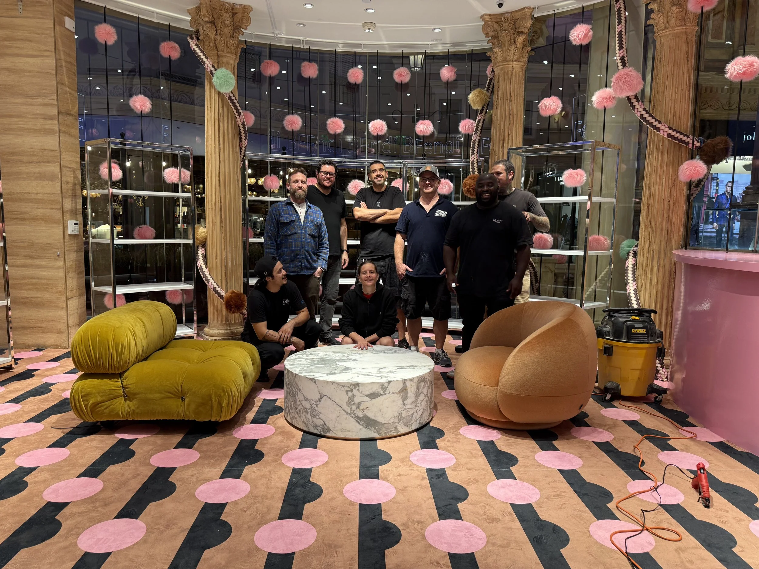 A group of seven people standing and sitting in a colorful, stylish store or lounge with pink and green decor. The background features large glass windows, pink pom-pom decorations hanging from the ceiling, and marble and velvet furniture.