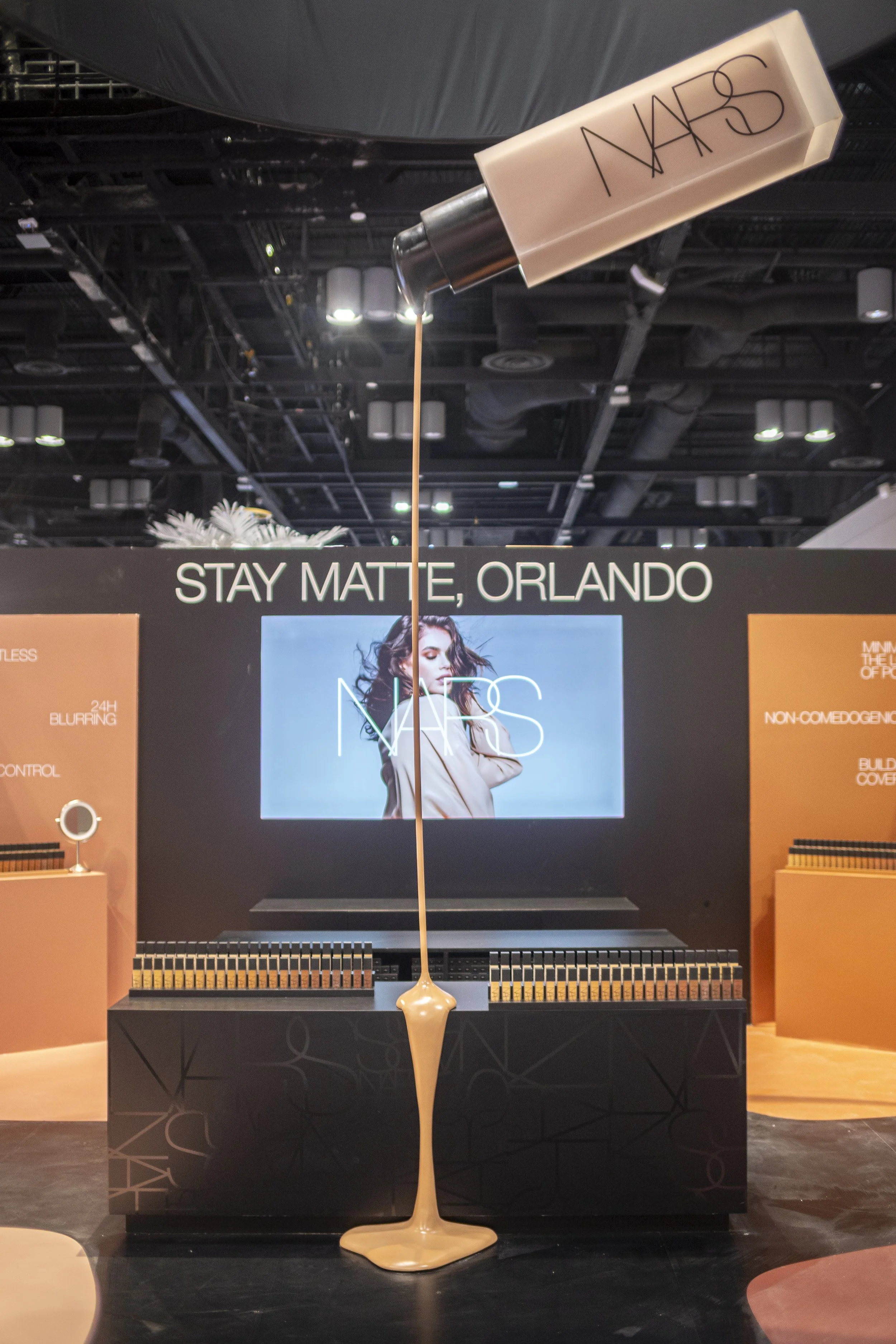 Display at a NARS cosmetics booth with a large bottle of foundation liquid coming from the bottle. There is a large screen showing a woman with makeup. The display has the text "STAY MATTE, ORLANDO" and NARS logo.