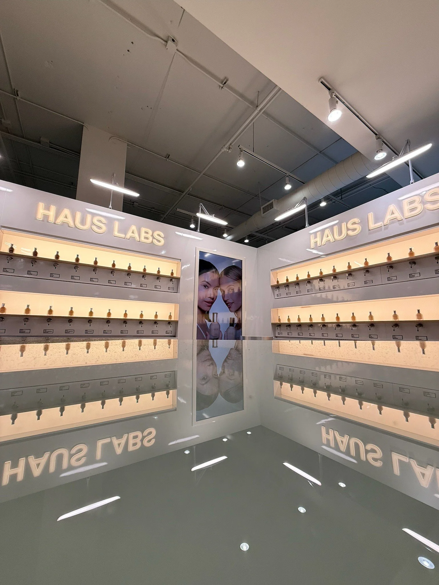 Interior of a store named HAUS LABS with illuminated signage, shelves displaying multiple small bottles, and a poster of two women holding perfume bottles.