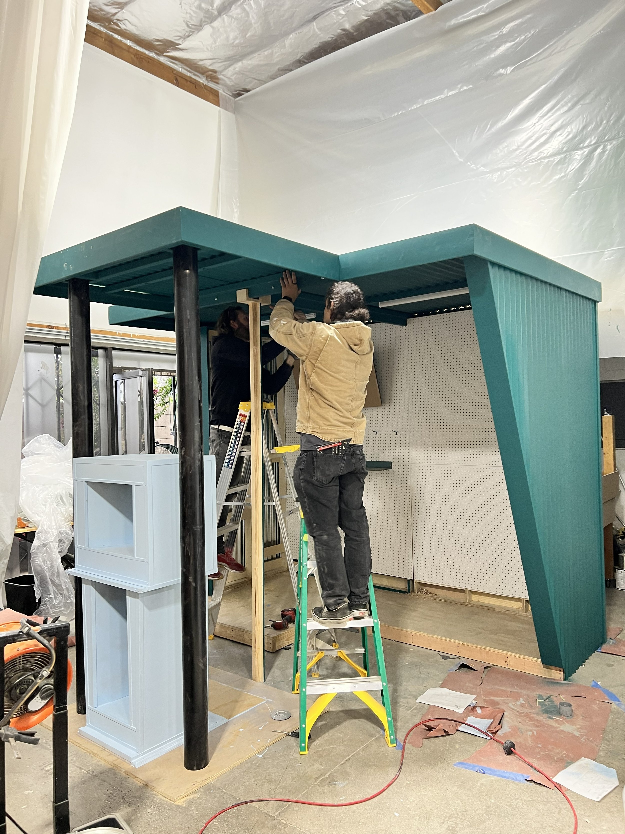Two people working on a construction project indoors, standing on a step ladder, attaching a vertical wooden beam to a teal-colored structure. The space has tools, materials, and construction debris around.