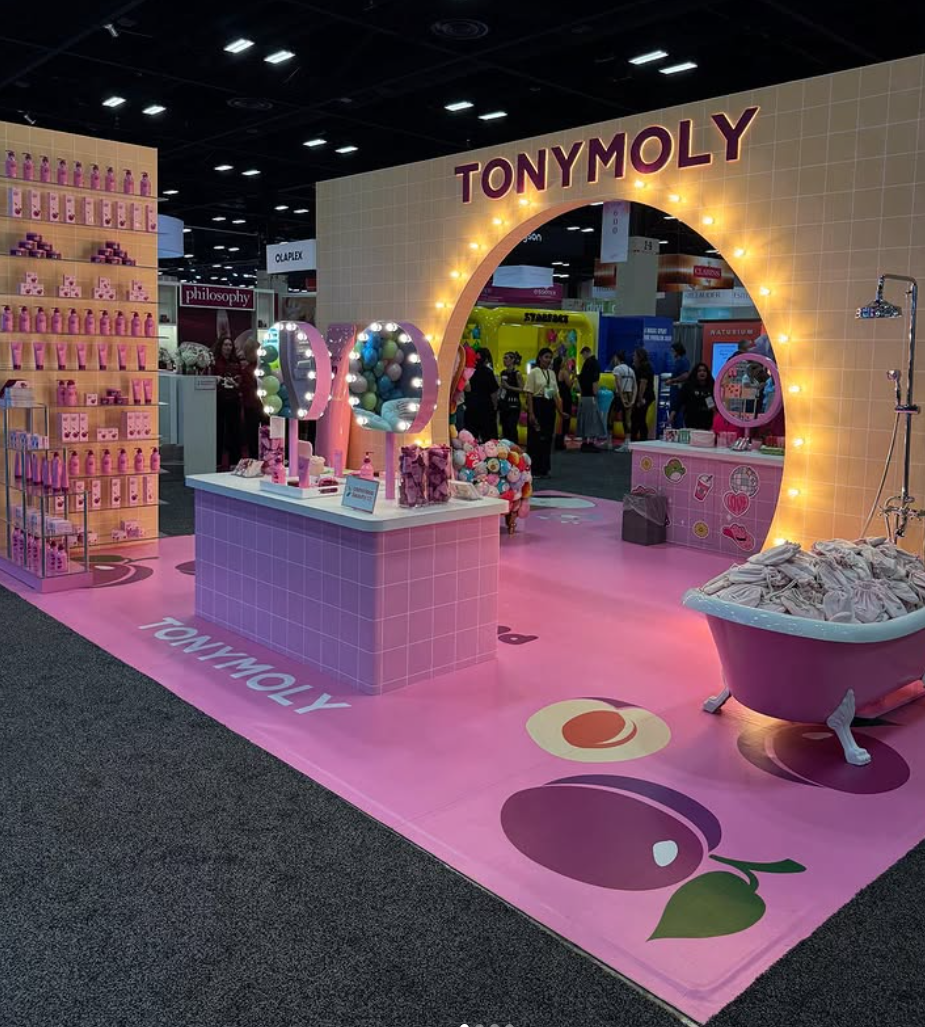 Pink-themed Tony Moly booth at an exhibition with a large archway, illuminated with string lights, and various cosmetics and skincare products displayed on shelves and counters. People are walking in the background.