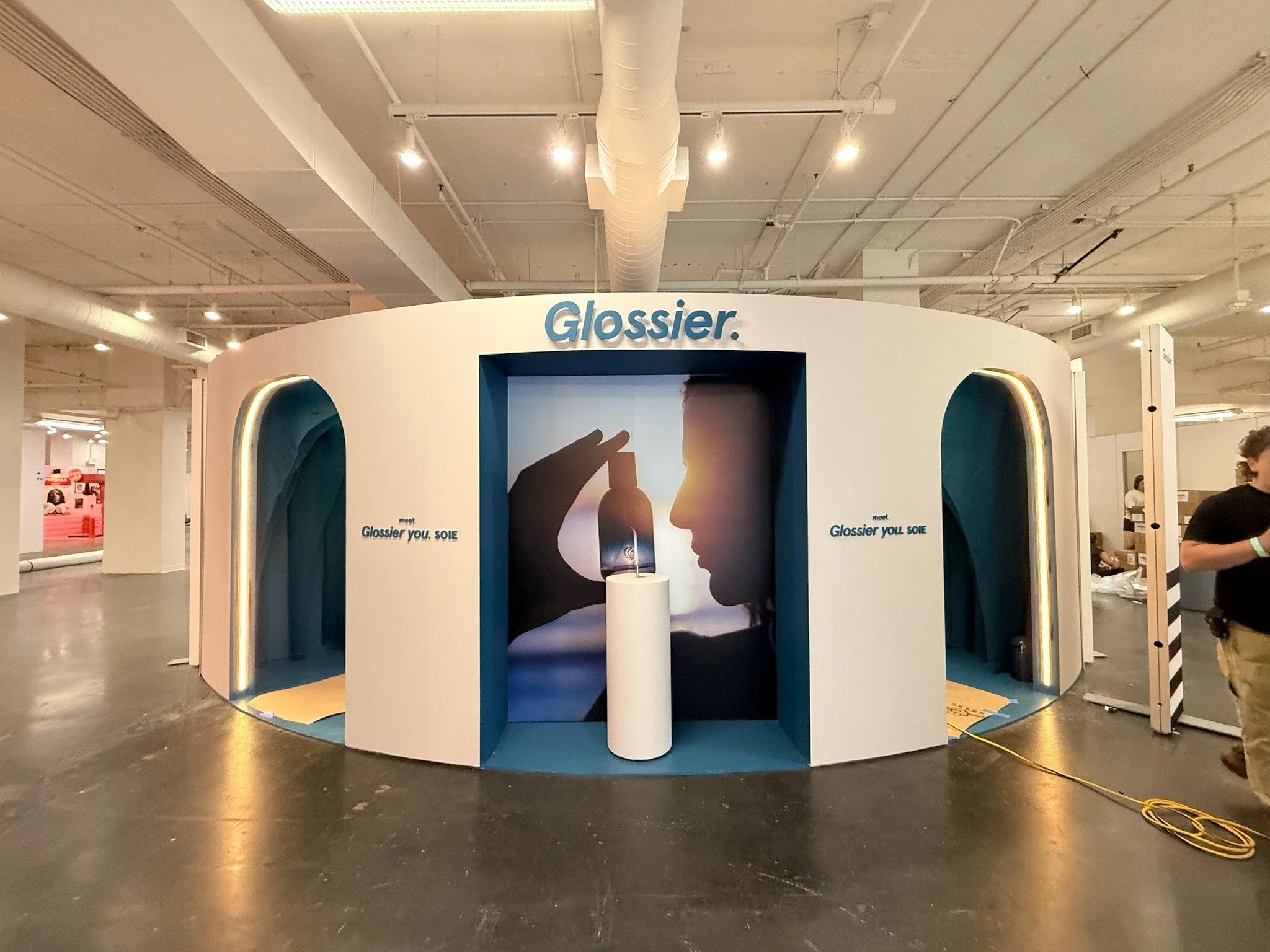 Booth with a sign that says 'Glossier' at an indoor event, featuring a graphic of a person holding a phone and a mirror.
