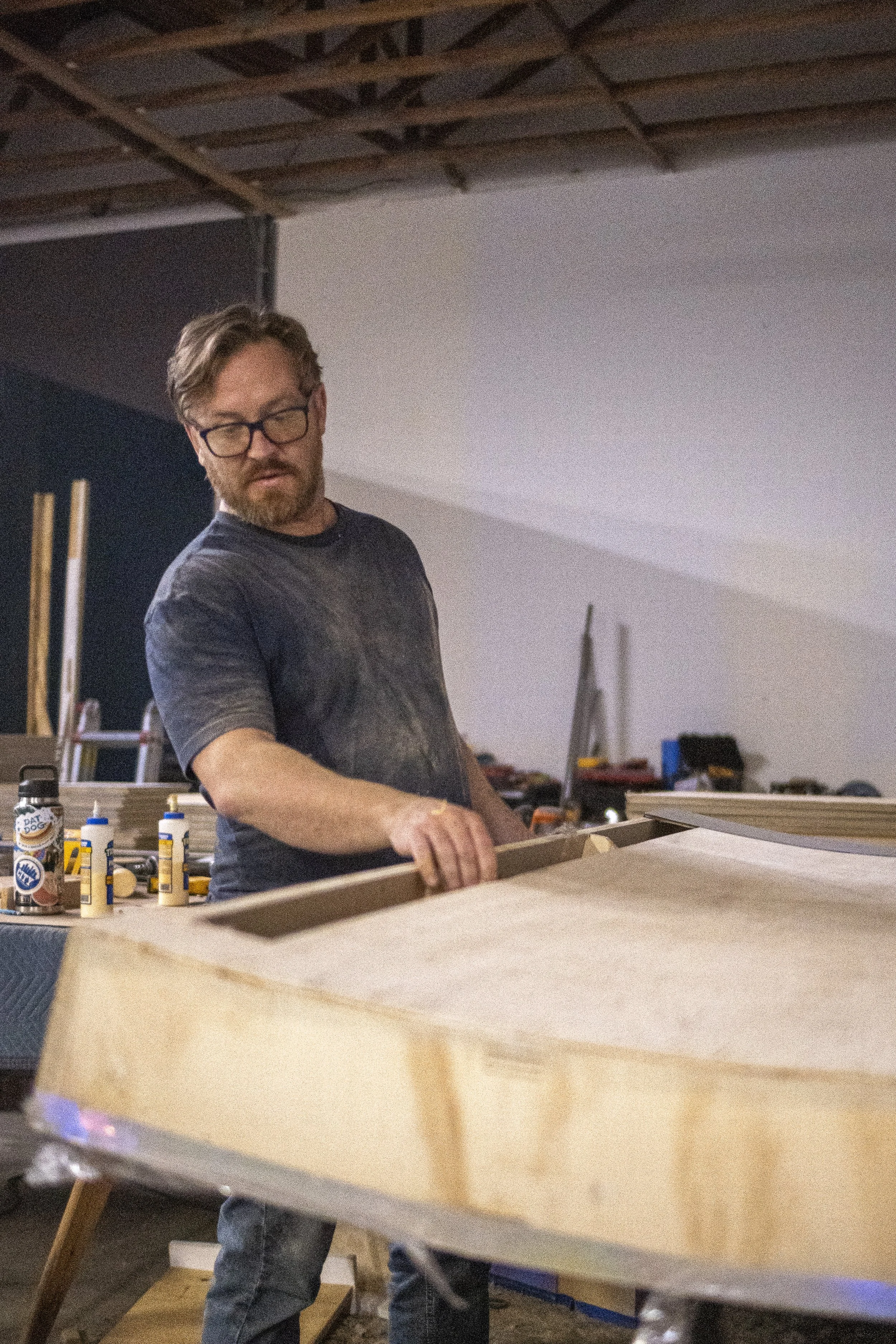 A man wearing glasses and a dark t-shirt is working on a wooden project in a workshop. There are bottles of glue and woodworking tools on the table. The background shows a partially unfinished ceiling and various workshop items.