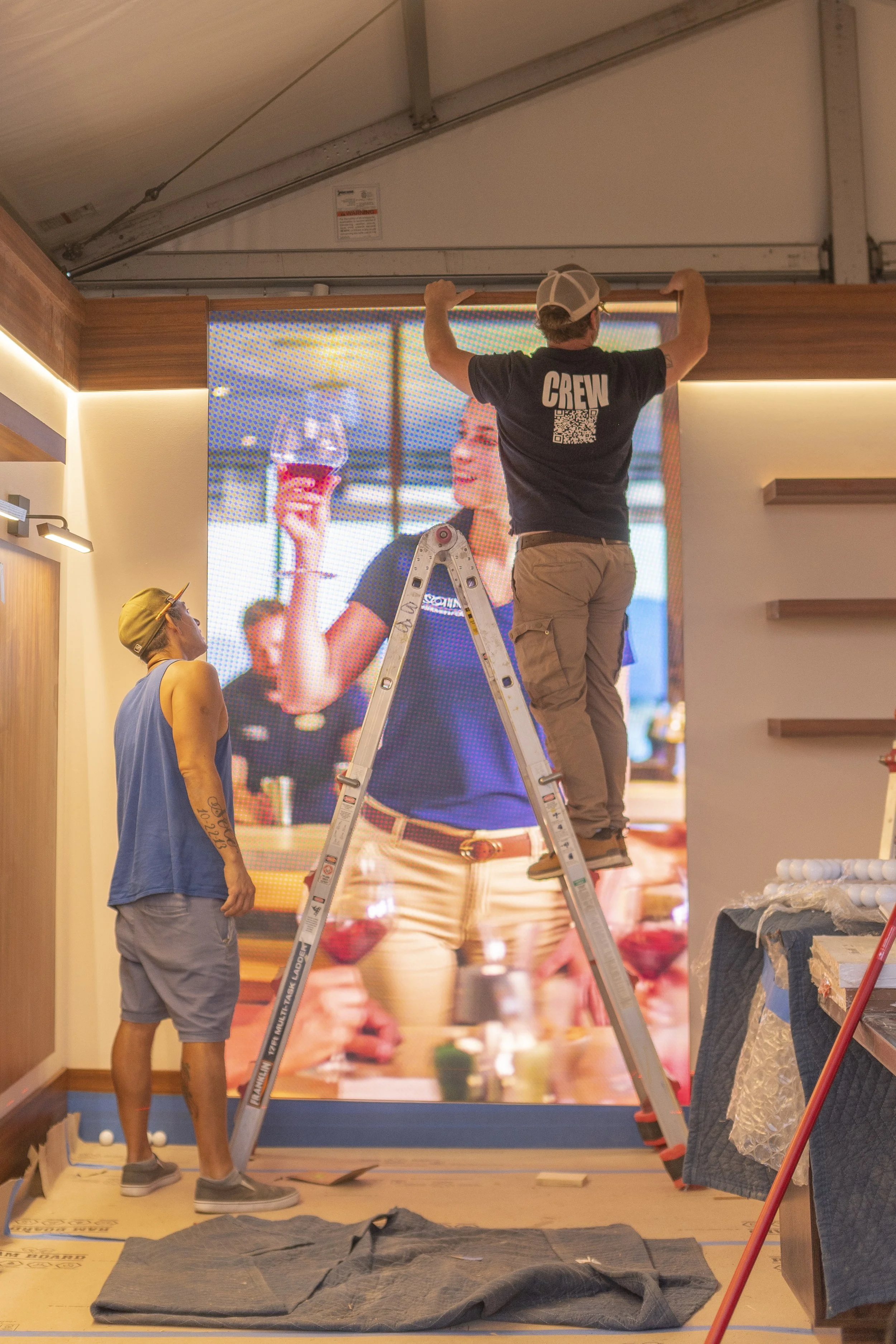 Workers installing a large LED display in a room with wooden accents. One worker stands on a ladder adjusting the top of the display, while another observes nearby. Construction materials and tools are visible on the floor.