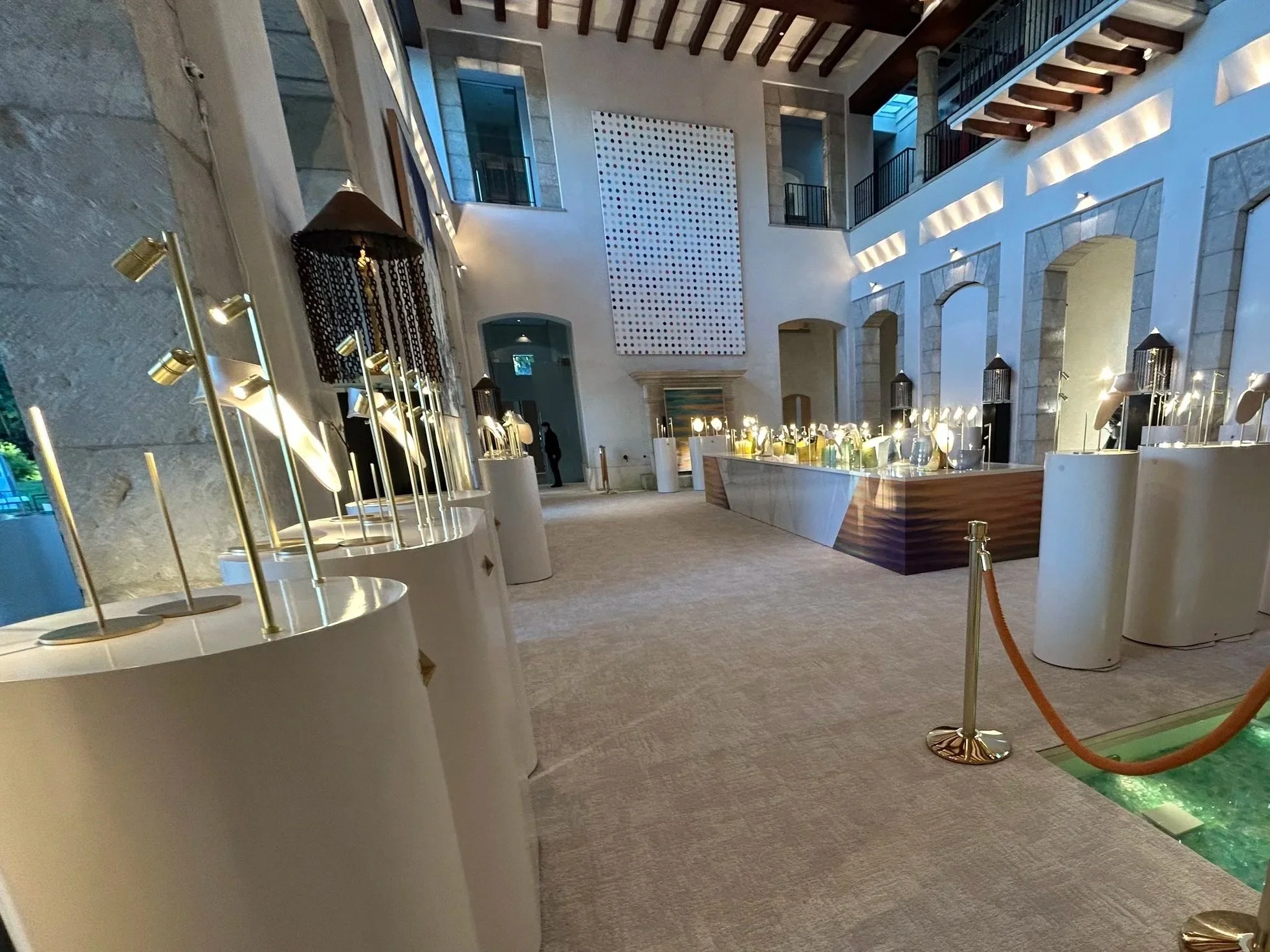 Interior view of a large hotel lobby or atrium with high ceilings, balconies, and modern lighting, featuring display tables with lamps and decorative items, and roped-off walkway area.