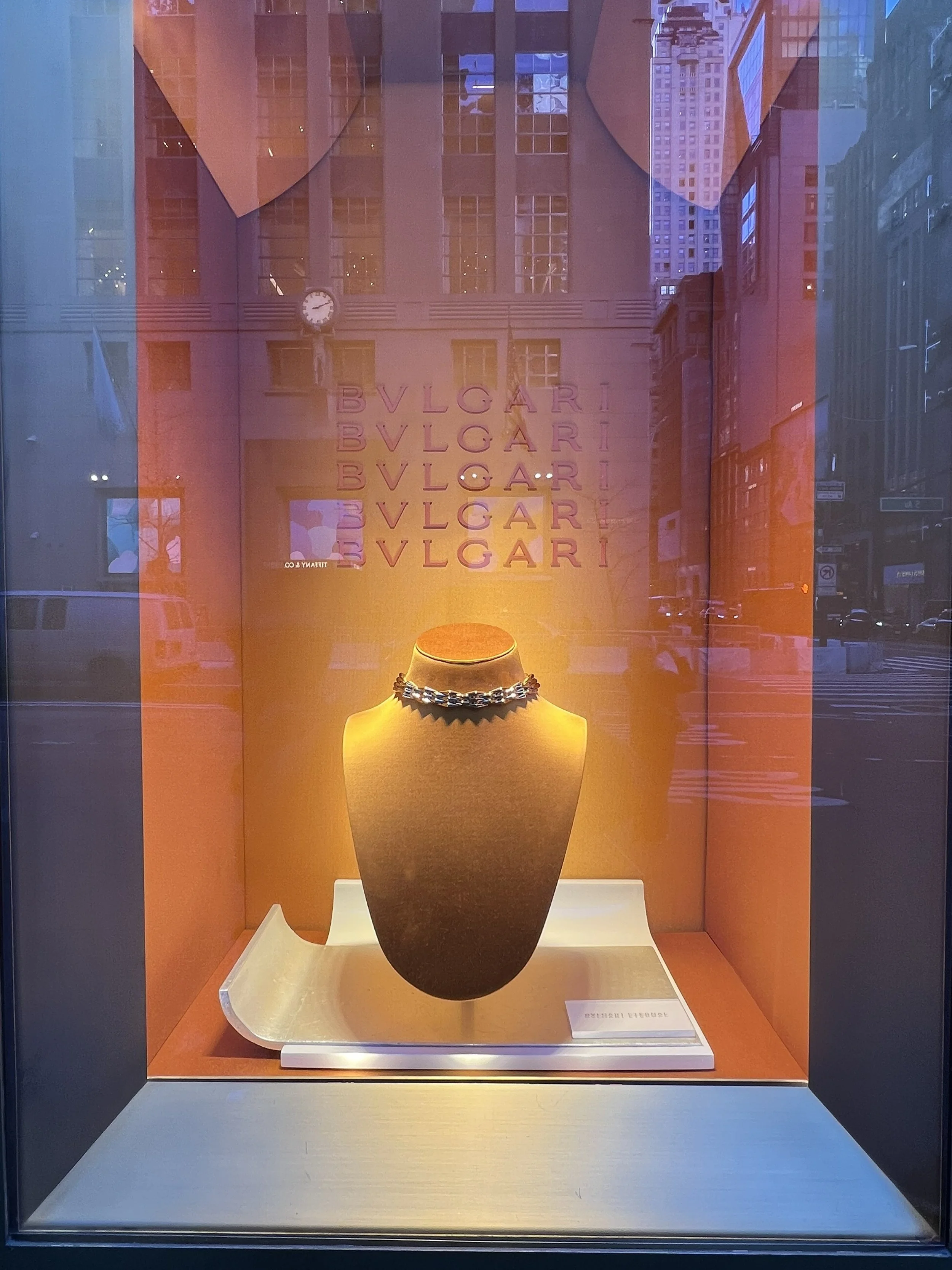 Jewelry displayed in a glass case with an orange interior background. The jewelry appears to be a necklace placed on a dark, rounded stand in the center of the display.