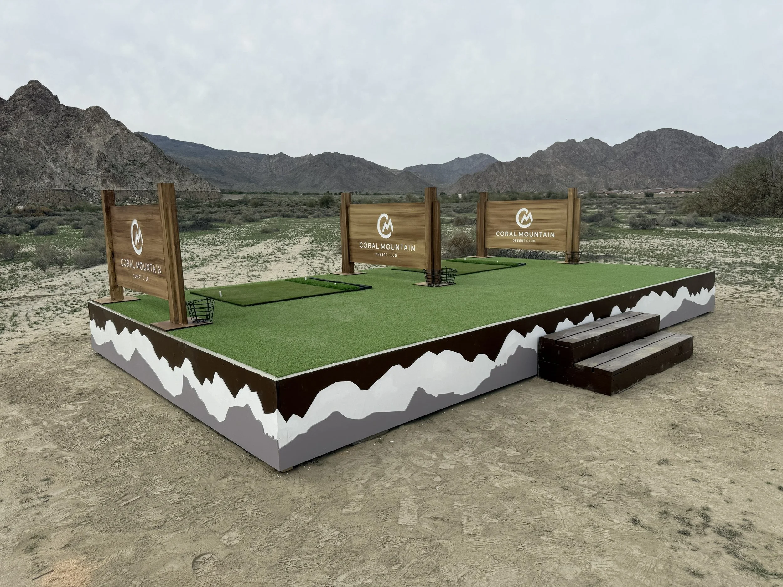 A golf driving range setup in a desert landscape with mountains in the background. The setup includes black mats and three wooden signs with the 'Coral Mountain' logo, surrounded by a white mountain range outline, placed on a platform with artificial