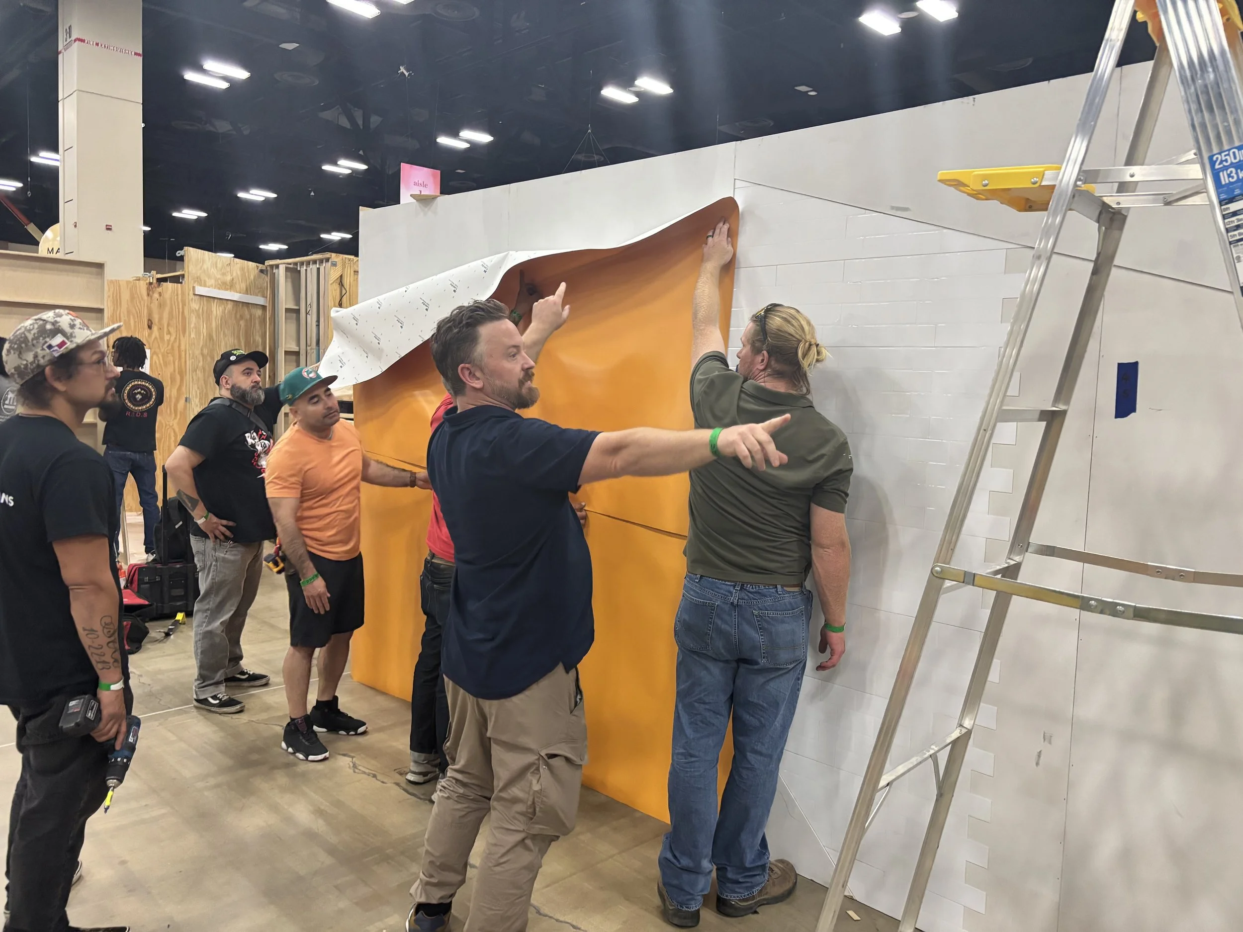 Group of people installing large orange wall panel in an indoor exhibit space, with ladder and construction tools present.