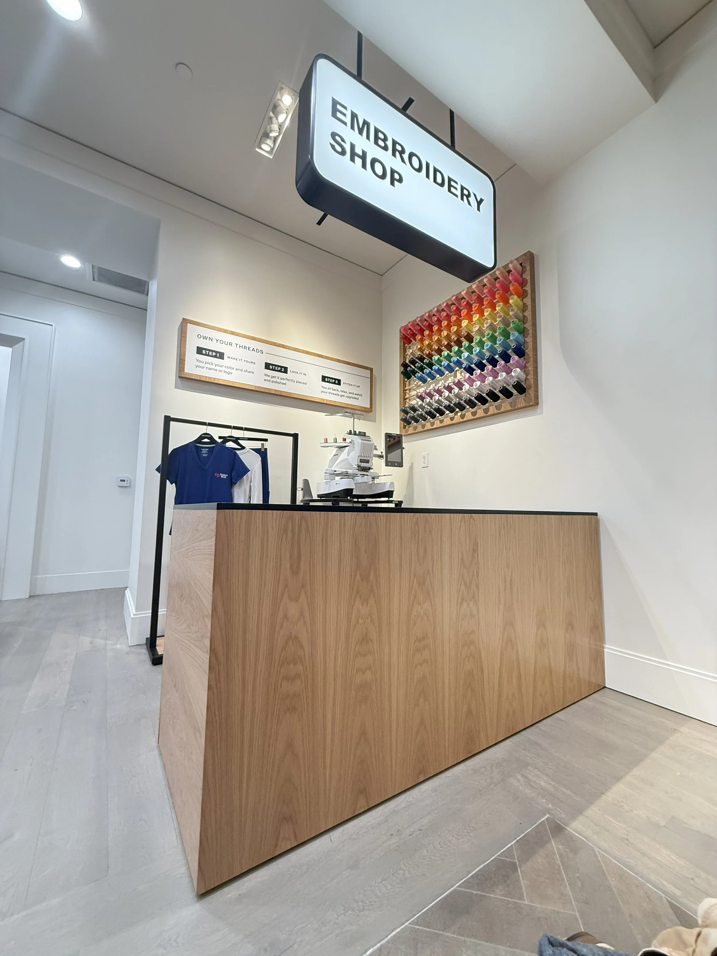 A retail store or shop with a sign reading 'Embroidery Shop' hanging from the ceiling. The store has a wooden counter with embroidery threads on the wall and a sewing machine behind the counter. There is a clothing rack with blue T-shirts nearby.