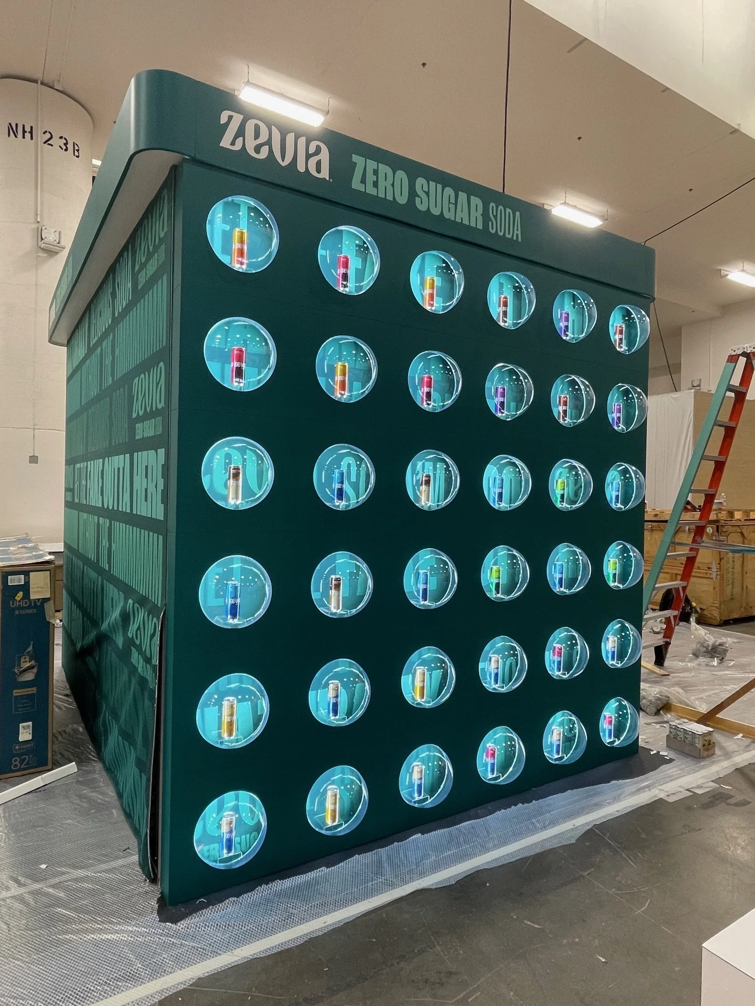 A green display stand with multiple circular windows showcasing cans of Zevia zero sugar soda in various flavors, with the Zevia logo and 'Zero Sugar Soda' written on top.