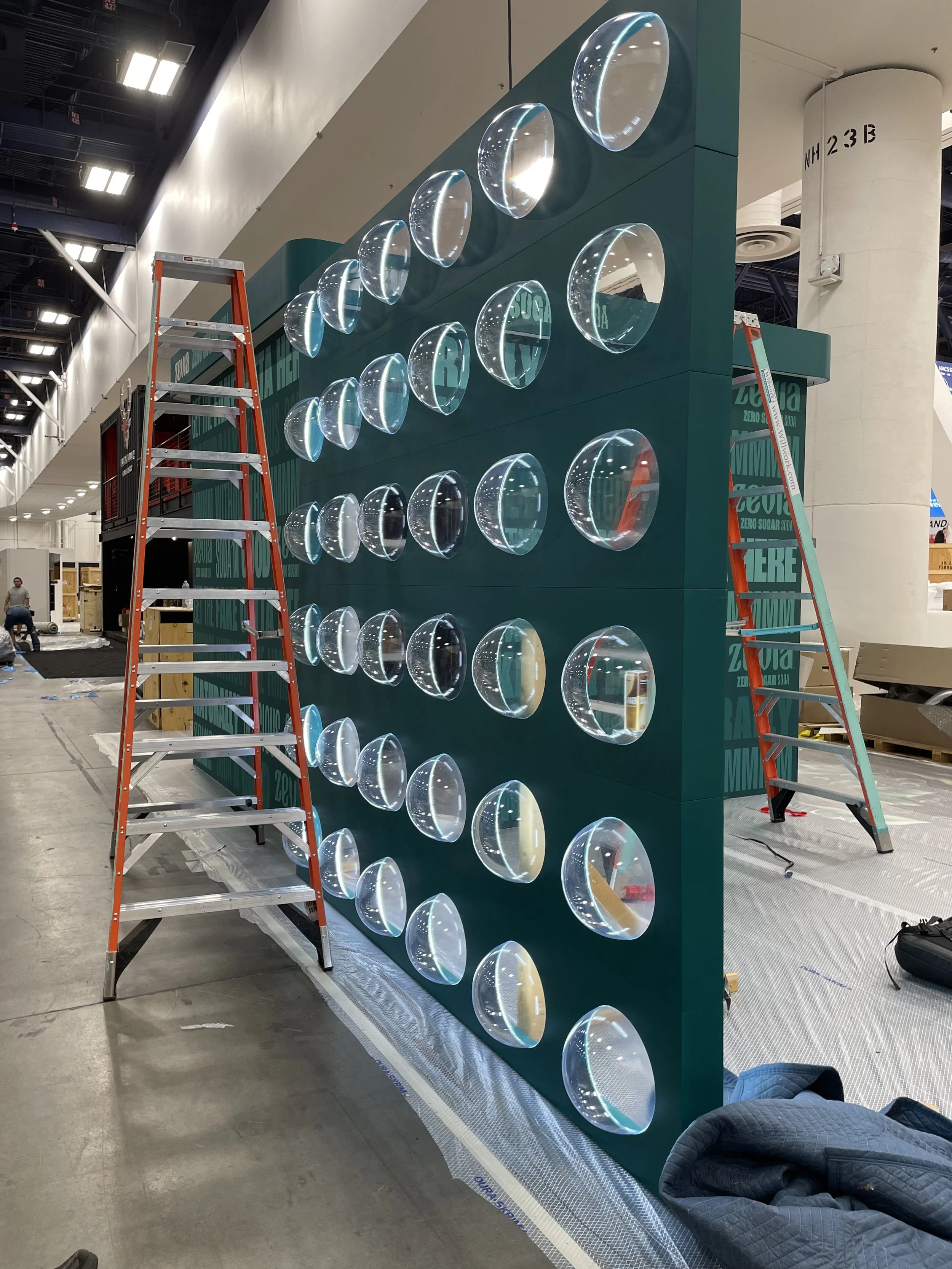 A large teal display with multiple clear, irregularly-shaped glass bowls or lenses mounted on it, situated in an indoor exhibition or convention space. Two ladders are positioned on either side of the display, possibly indicating ongoing installation