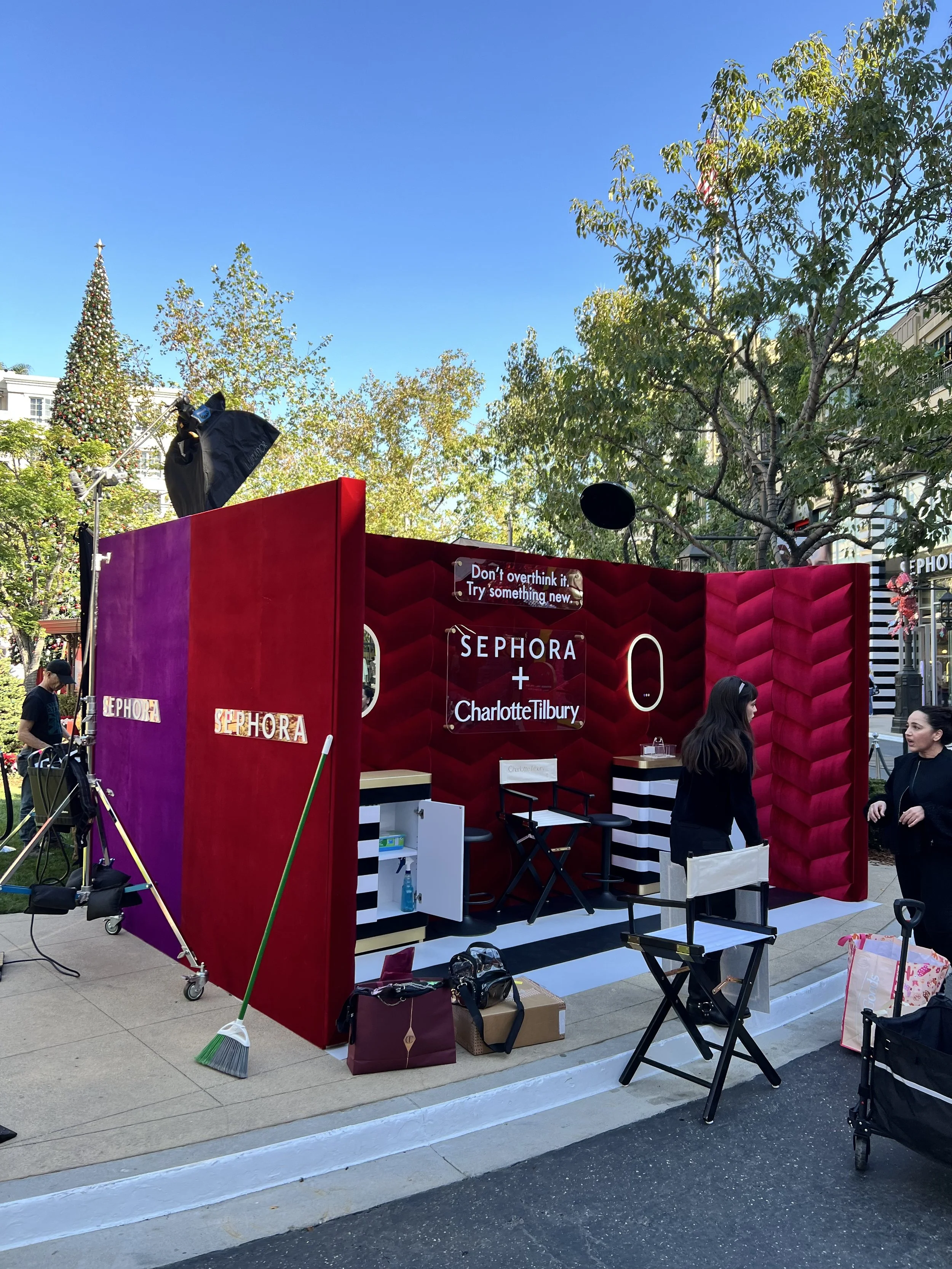 Outdoor Sephora pop-up booth with red and black decor, sign featuring names Charlotte Tilbury and Sephora, surrounded by trees, people, and city buildings on a sunny day.