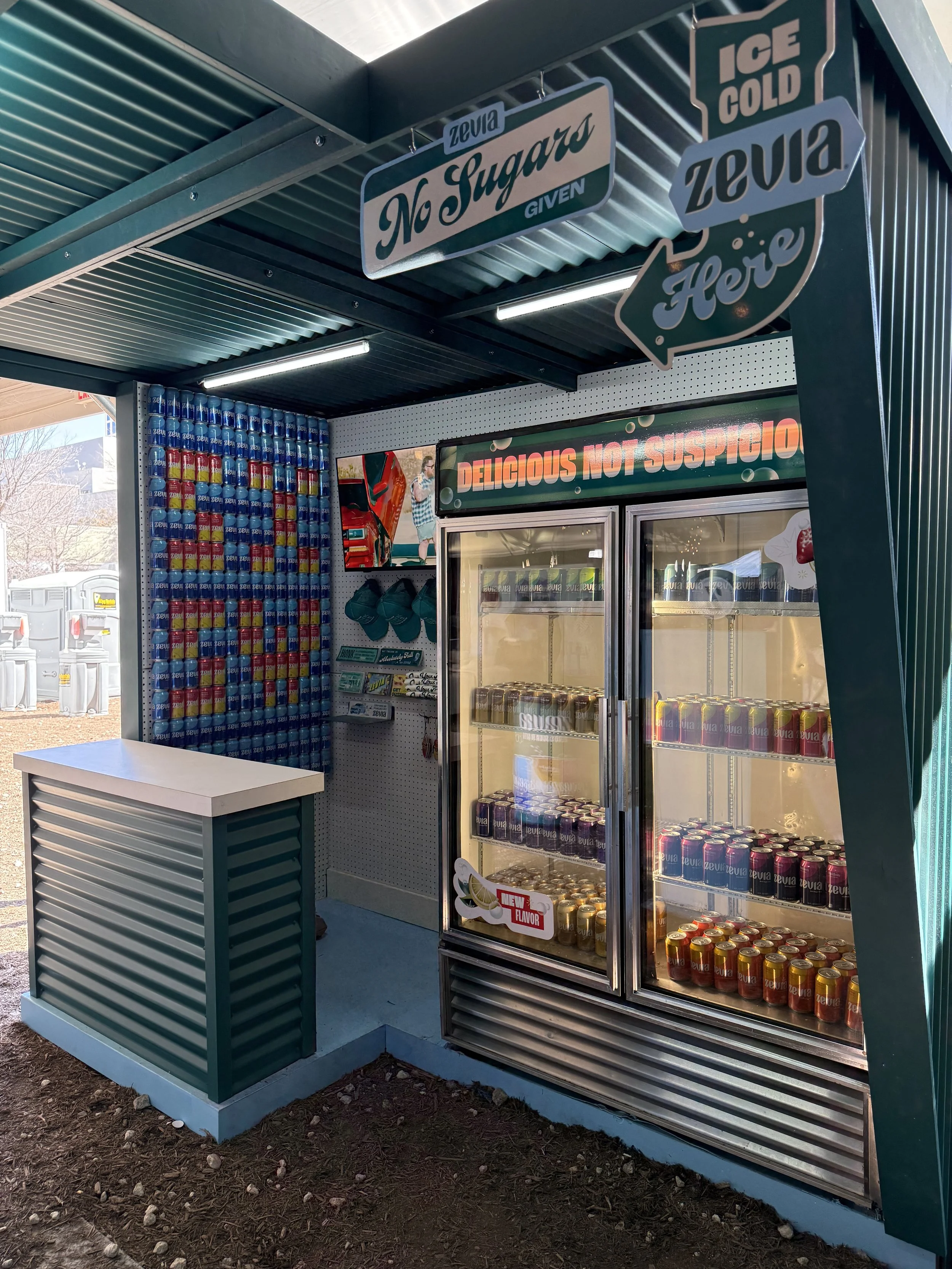 A vending booth with a glass fridge displaying cans of Zevia soda, with signs that say "No Sugar," "Ice Cold," and "Delicious Not Suspicious."