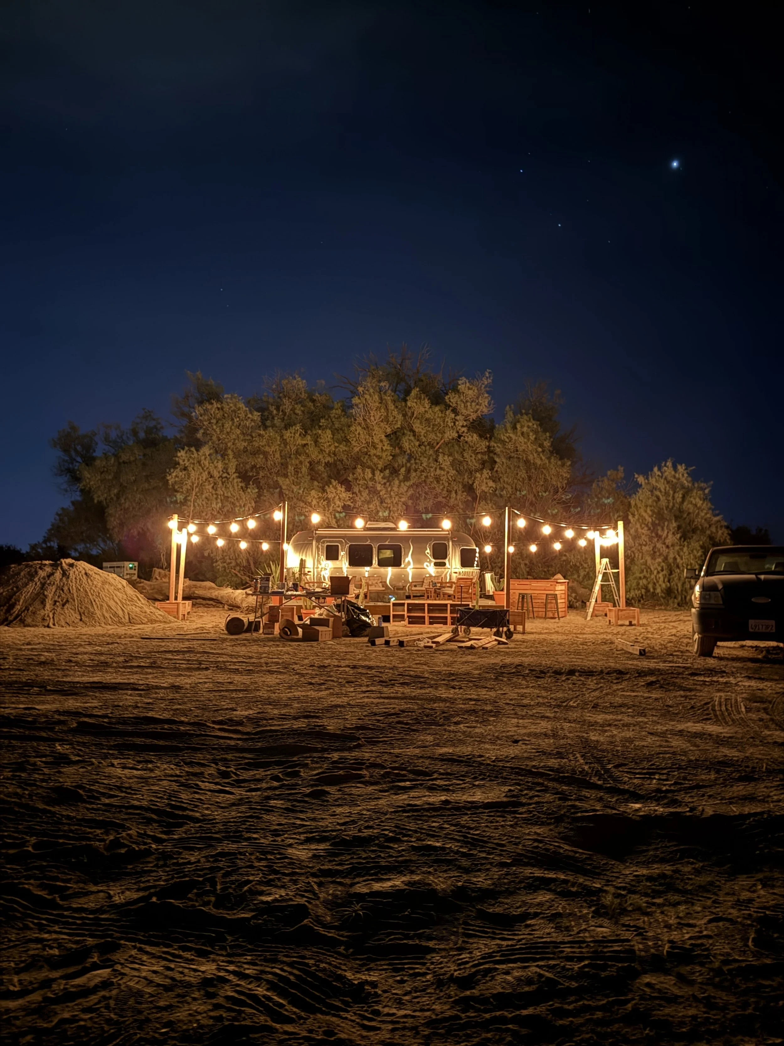 Nighttime outdoor scene with string lights illuminating a setup including a small trailer, tables, chairs, and various objects, with trees and a dark sky with stars in the background.