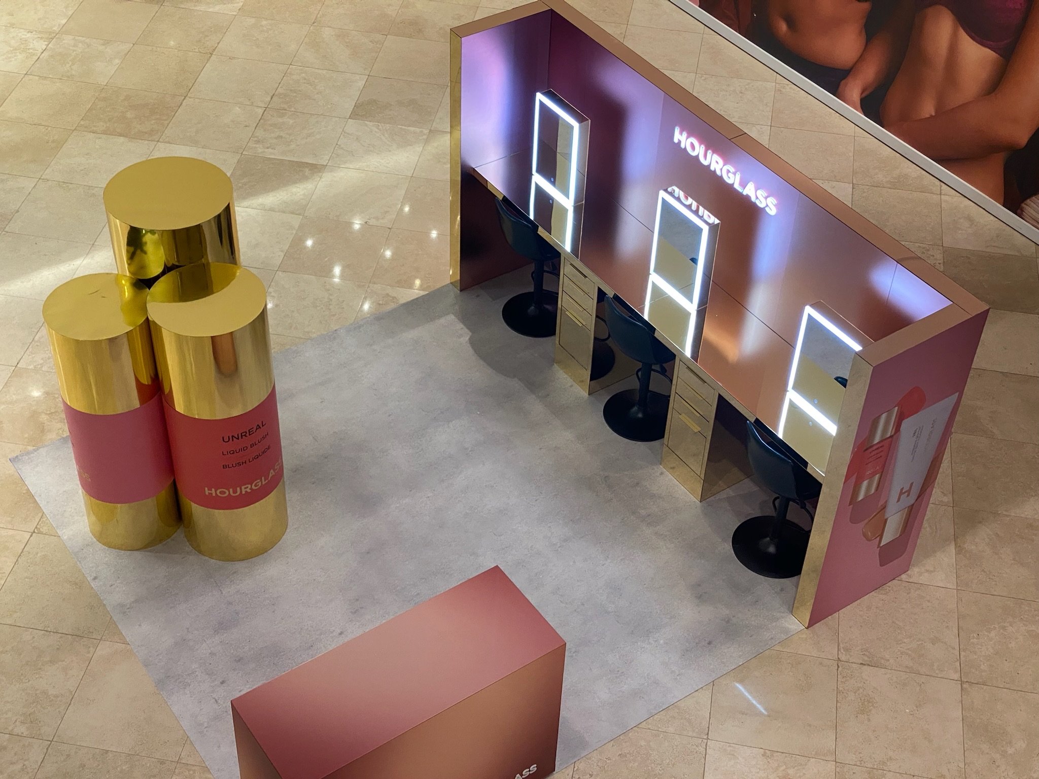 Decorative display with three gold-colored cylindrical containers stacked on top of each other on a pink base, and a modern stylistic reception or booth with illuminated rectangular frames, chairs, and signage for 'Hourglass' in a shopping mall or si