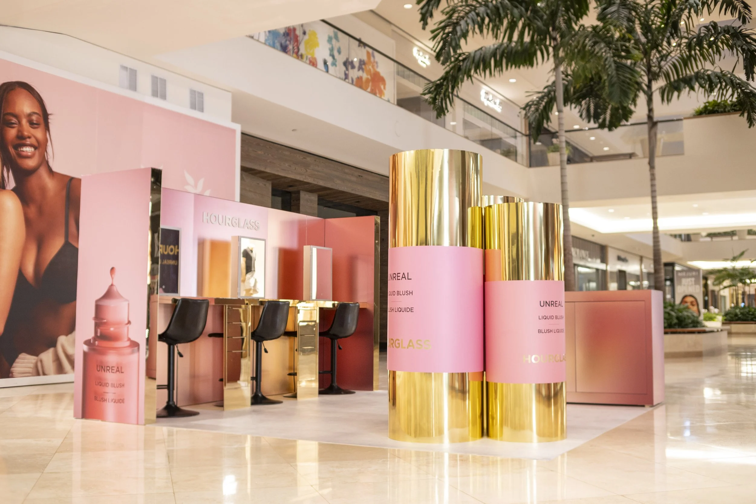 Display booth for Hourglass cosmetics in a shopping mall, featuring pink and gold themed products and signage, with a large poster of a smiling woman and some trees in the background.