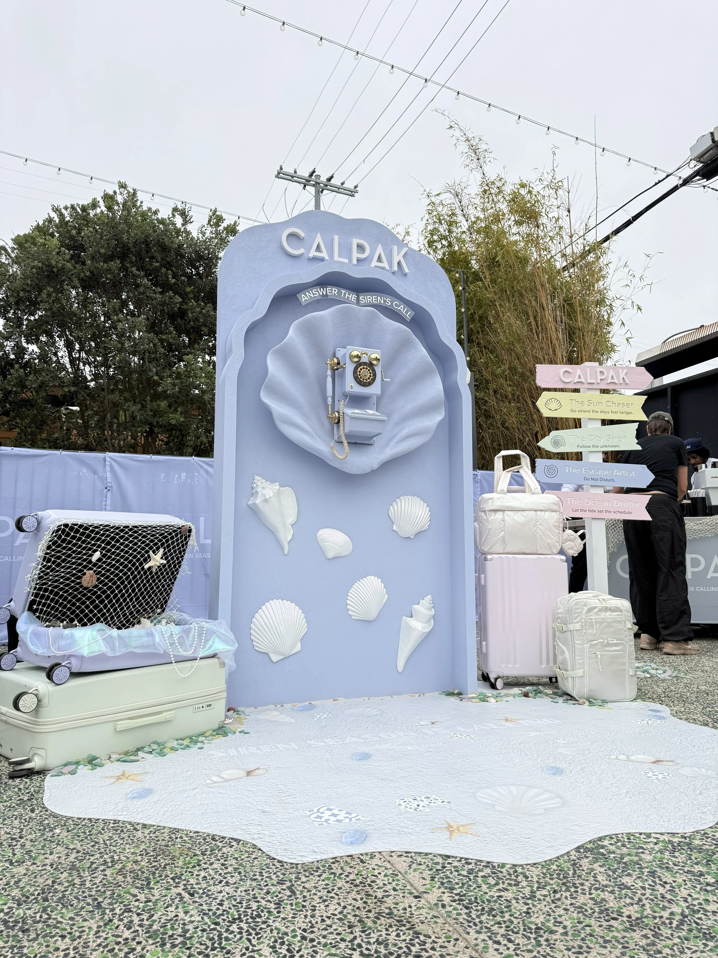 A decorative outdoor display with a light blue backdrop featuring a vintage rotary telephone and white seashell accents. There are black and white suitcases, some with jewelry and the floor is decorated with seashell and starfish designs, suggesting a beach or ocean theme.