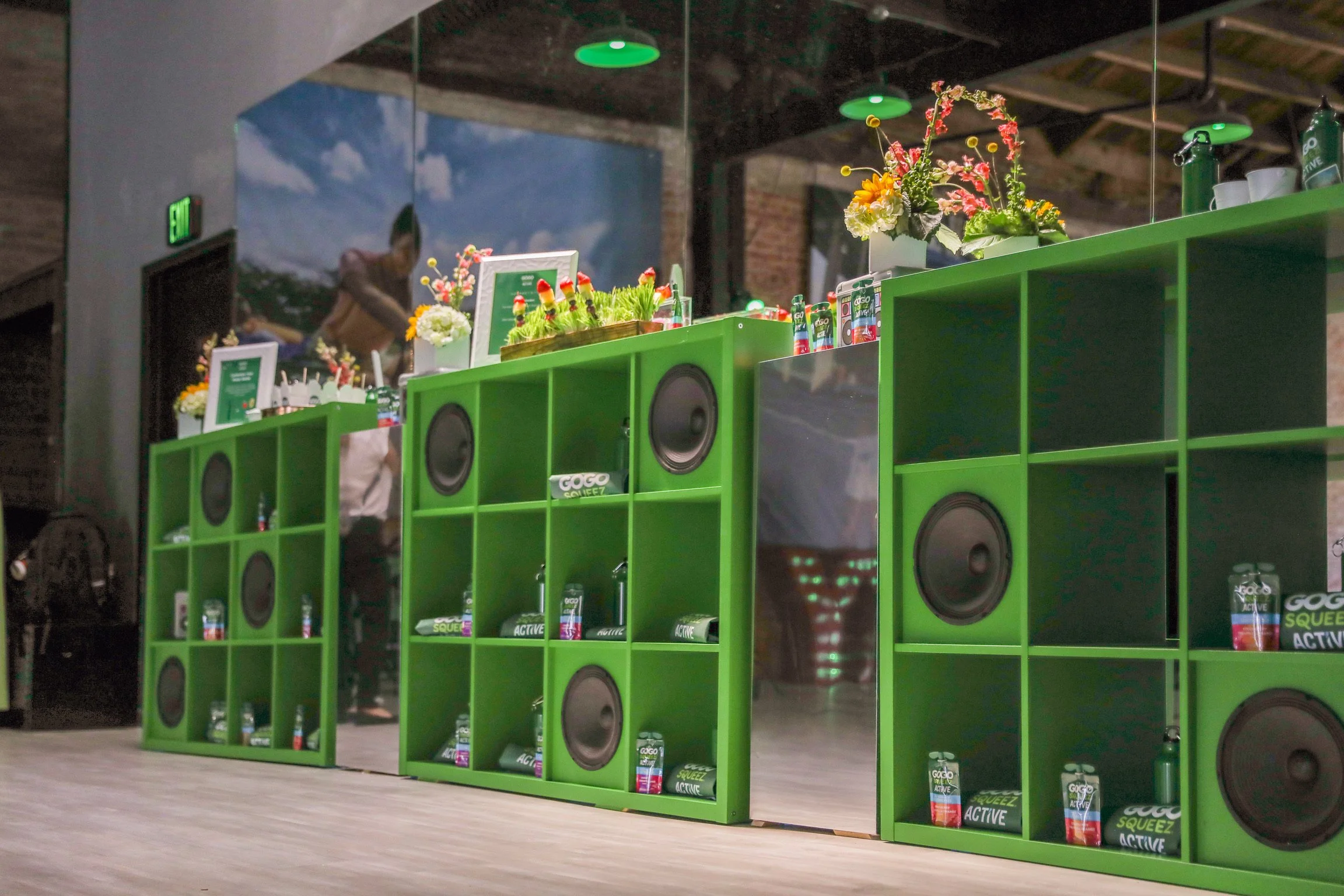 Green display shelves with gadgets and products, decorated with flowers, in an indoor space with a person in the background and wooden ceiling.