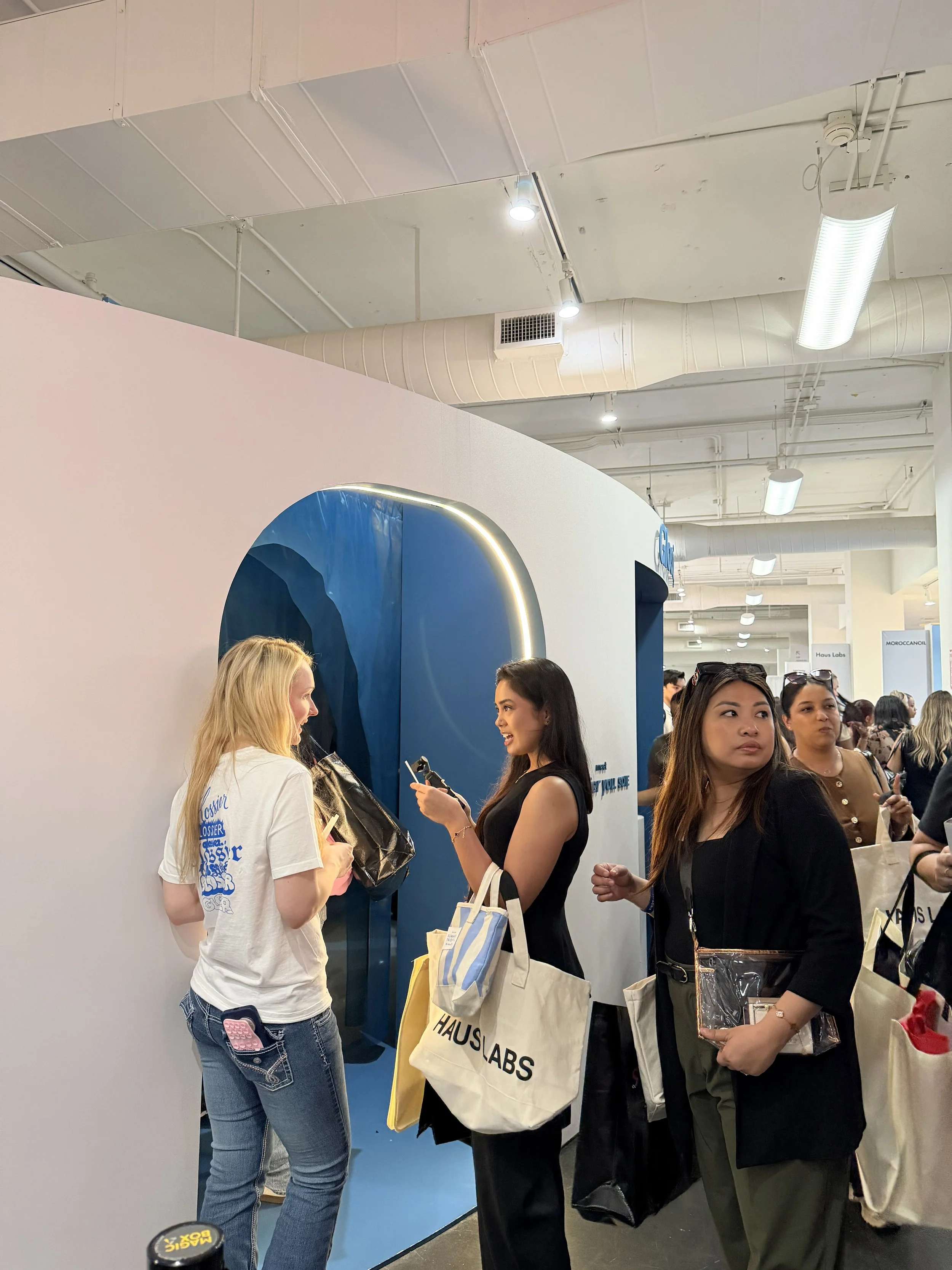 People standing in line at a busy indoor event, some engaged in conversation, with a booth or display in the background and shopping bags in hand.