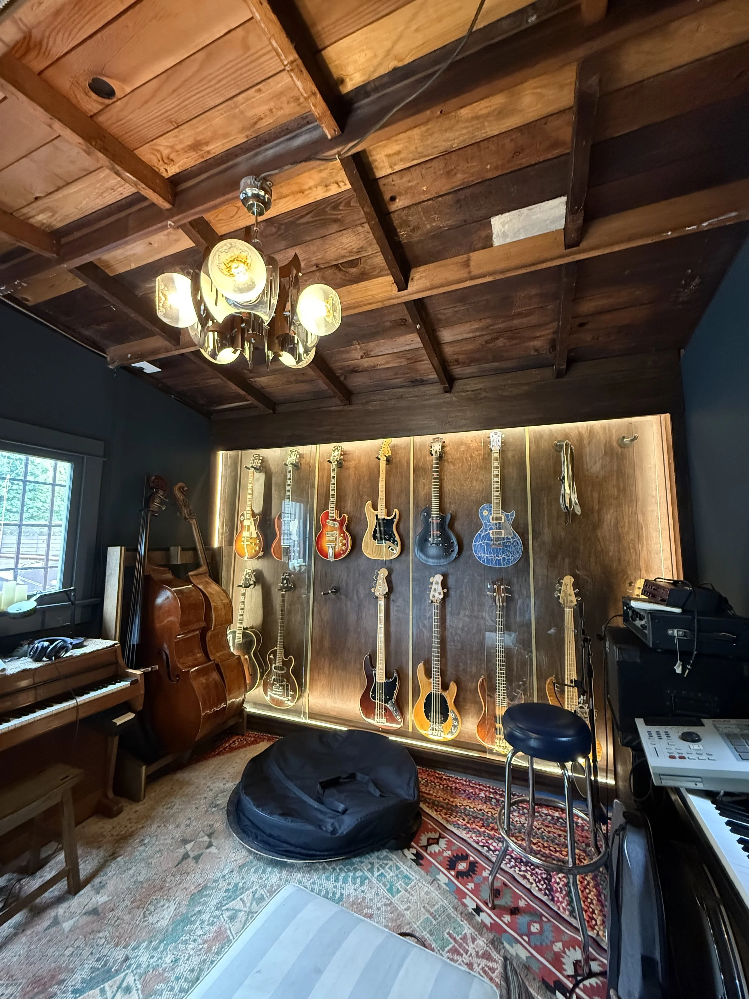 A music studio room with a collection of guitars displayed on a wooden wall, a black bean bag chair in the center, a piano on the left, and music equipment on the right, with a chandelier hanging from the wooden ceiling and a window on the left side.
