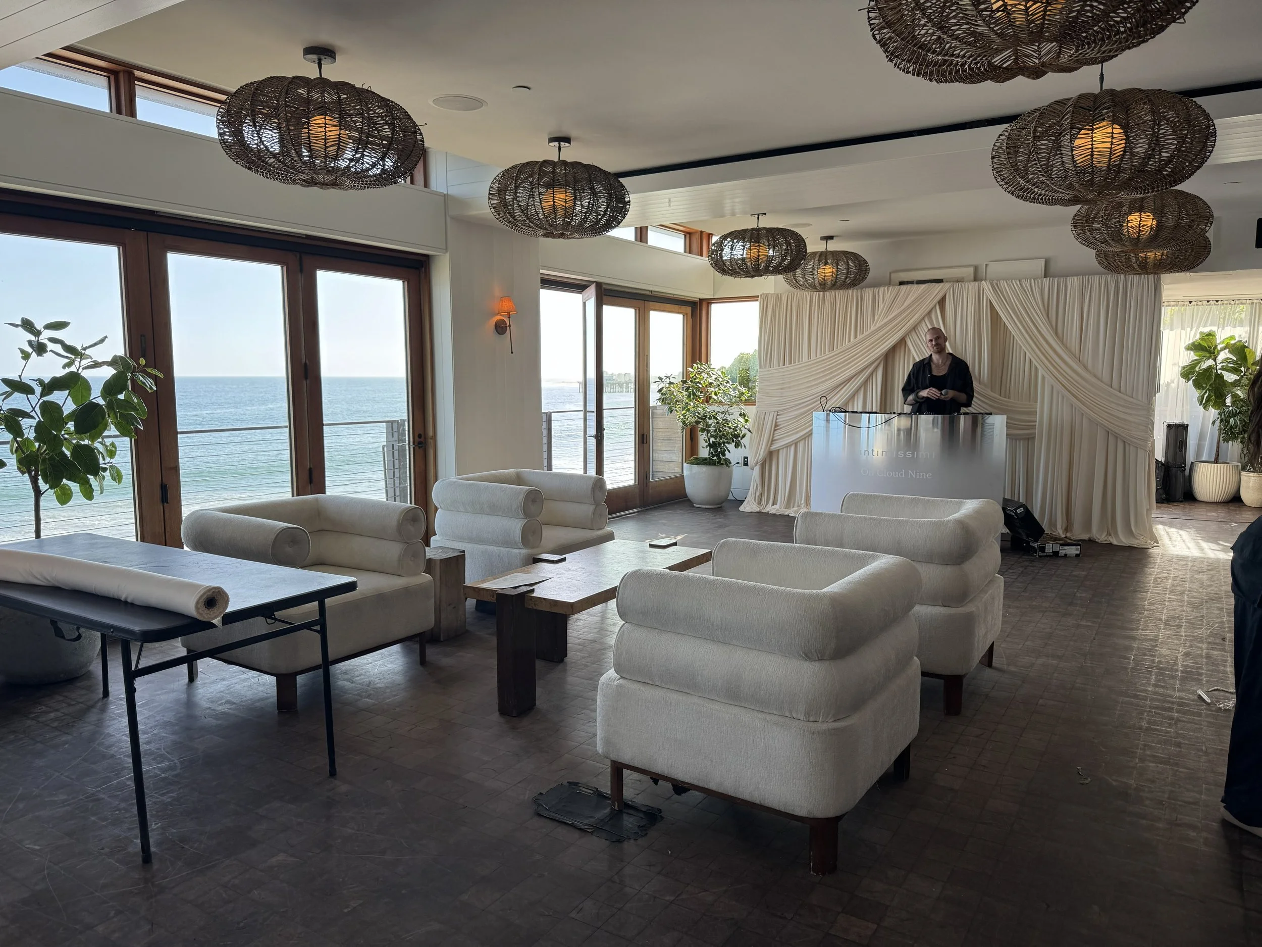 Interior of a modern room with large windows showing an ocean view, white furniture, chandeliers hanging from the ceiling, and a person standing at a DJ booth with a draped backdrop.