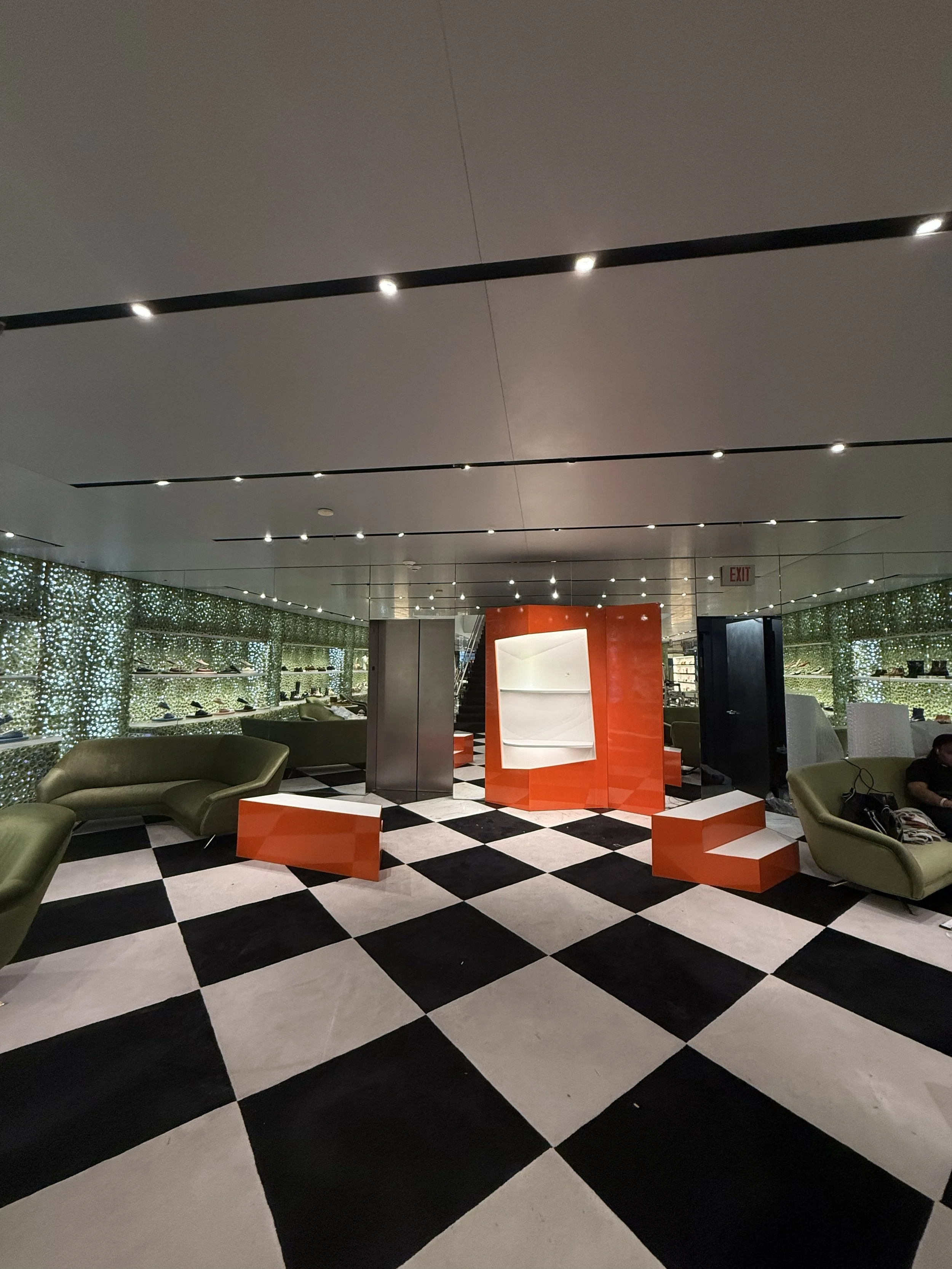 Modern interior space with black and white checkered floor, green armchairs, and orange geometric furniture, illuminated ceiling lights, and decorative wall panels.