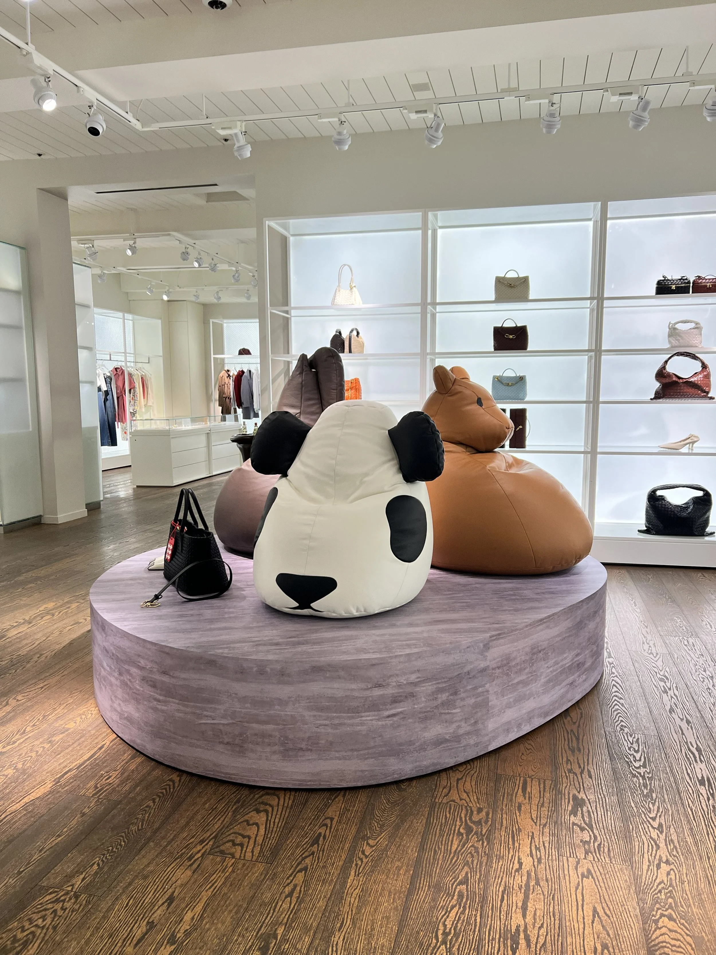 Display of handbags on white shelves in a retail store, with bean bag chairs shaped like dogs in front, situated on a circular platform with dark wood floor.