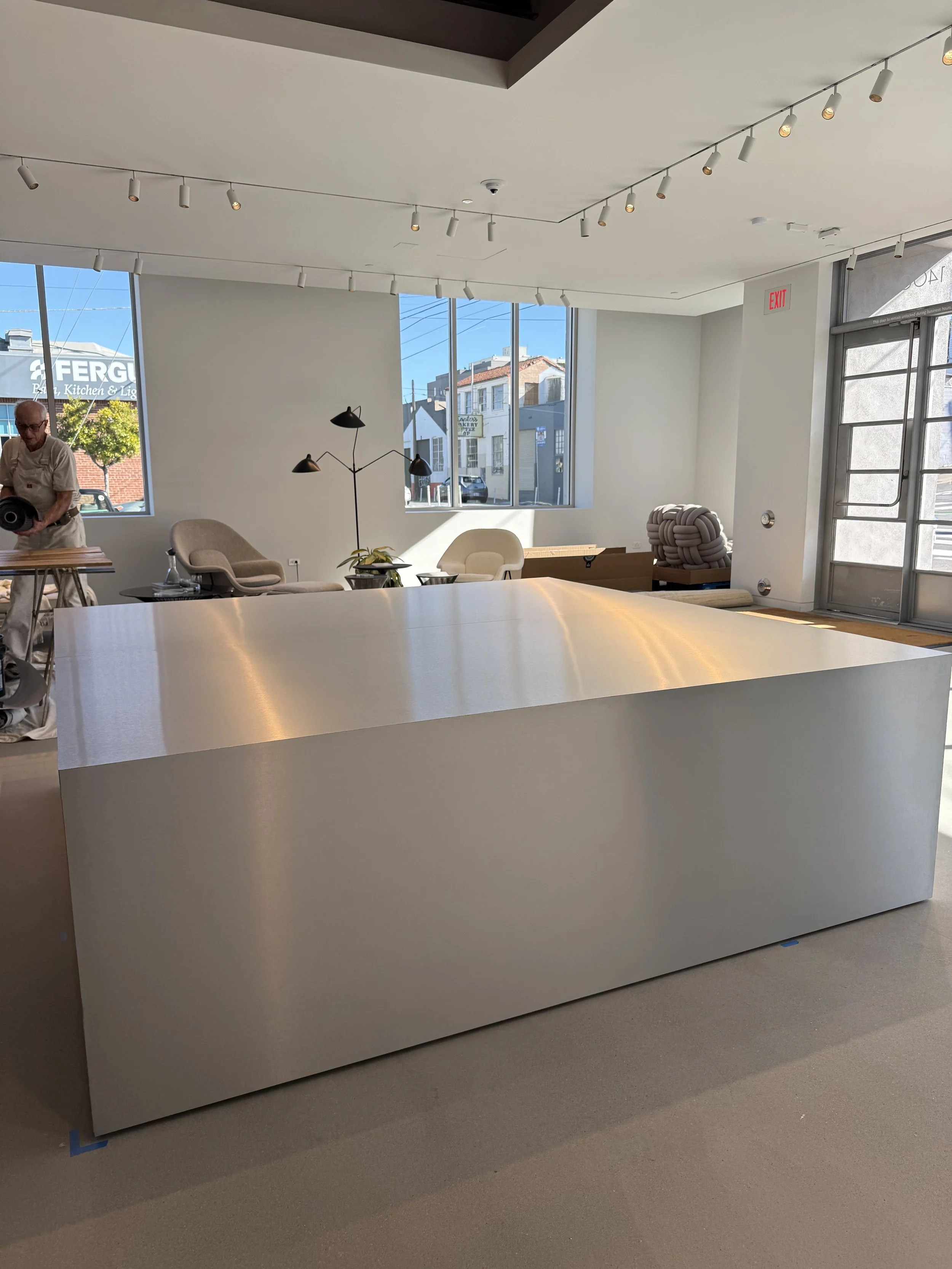 Interior of a modern commercial space with large windows, a white curved reception desk, lounge chairs, a lamp, and a potted plant, during daytime.