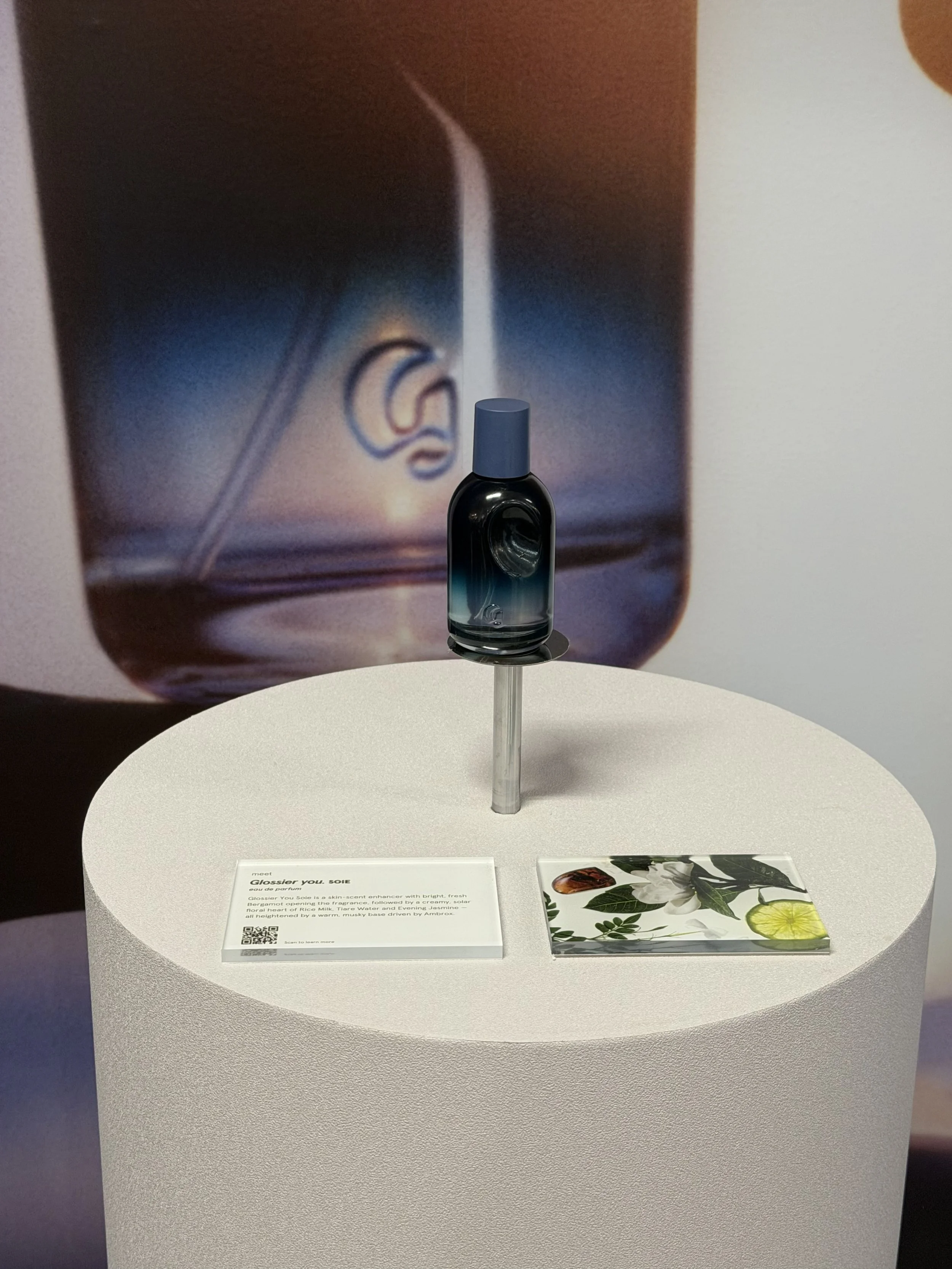 A small black perfume bottle with a blue cap on a round white display table. Behind it, there is a large poster featuring an ear in a field of blue and green hues, with text explaining a fragrance called 'GLOSSIER YOU, SOIE' and a QR code.