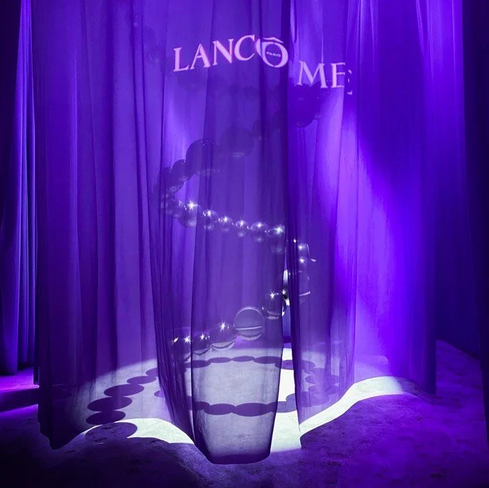 Purple-lit curtain with the words "LANCÔME" at the top, partially revealing a woman with jewelry behind it, creating a shadowed silhouette.