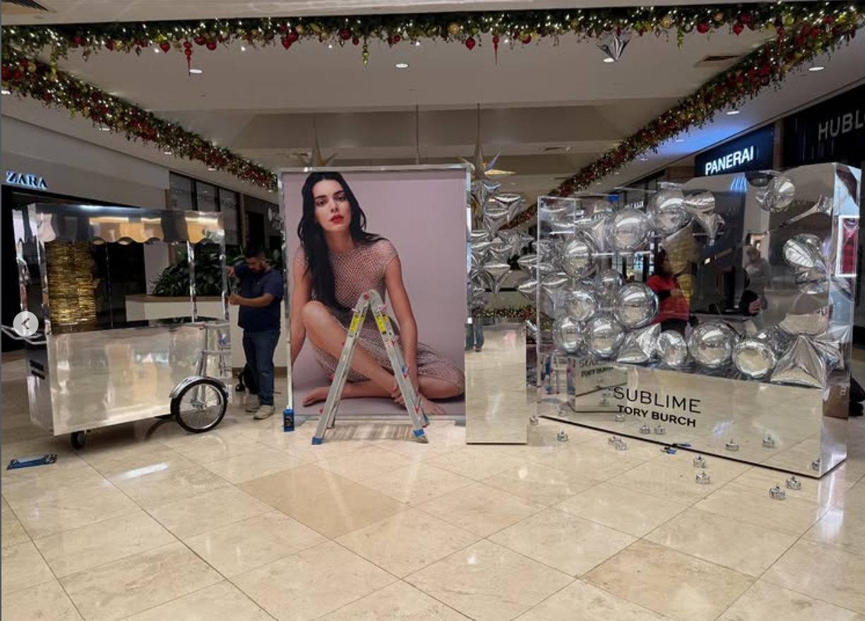 Christmas decorations and promotional displays inside a shopping mall, including a large photo of a woman, shiny balloon arrangements, and signs for watches like Panerai and Hublot.