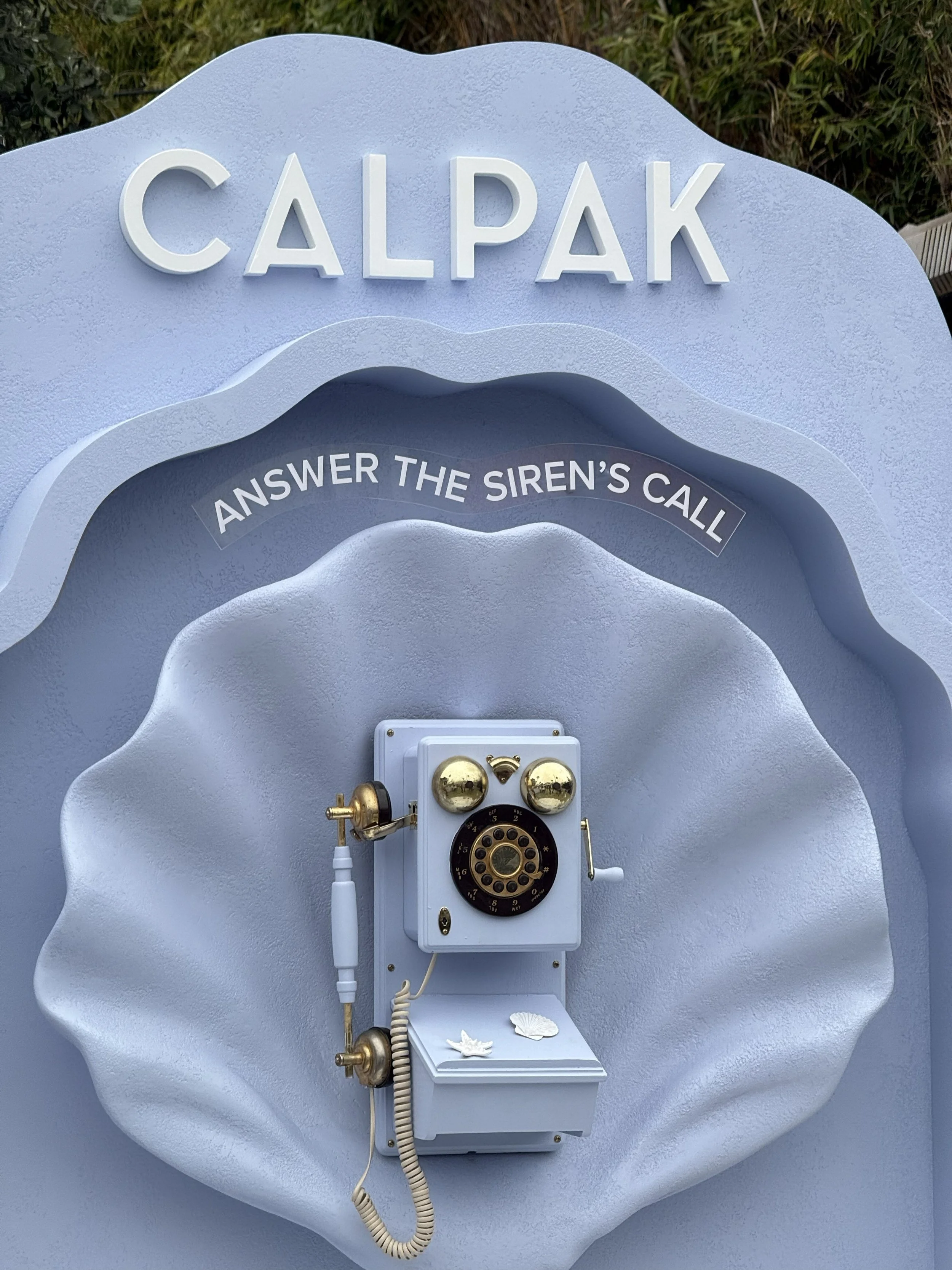 A decorative white surfboard-shaped display with a vintage telephone attached, featuring the text 'Calpak' and 'Answer the Siren's Call'.