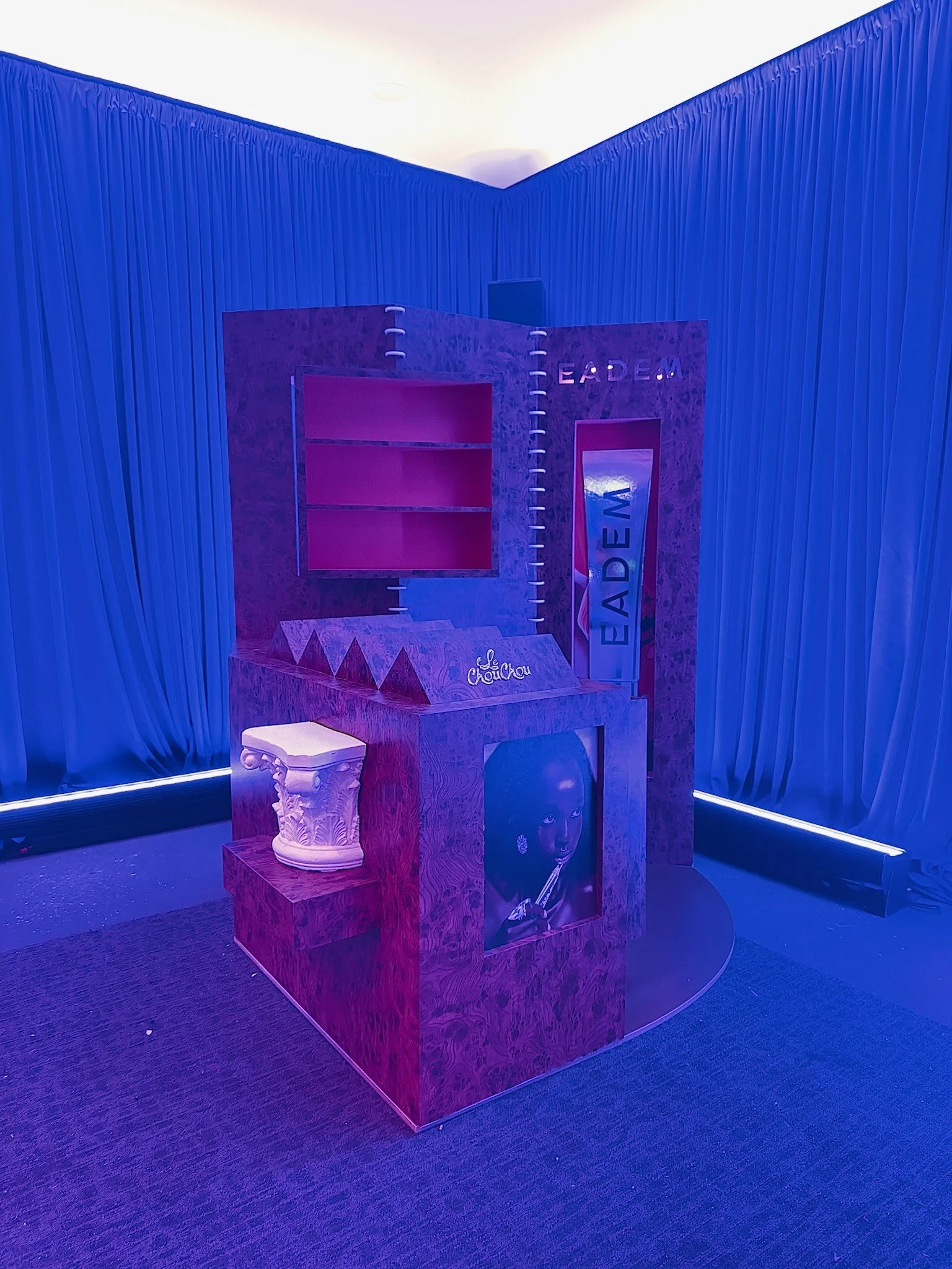 An art installation with a wooden structure featuring a picture of a woman with earrings and a cigarette, a top hat, and the words 'LEADER' and 'EAD' illuminated in purple and pink lights. The background has blue pleated fabric and a curved section w