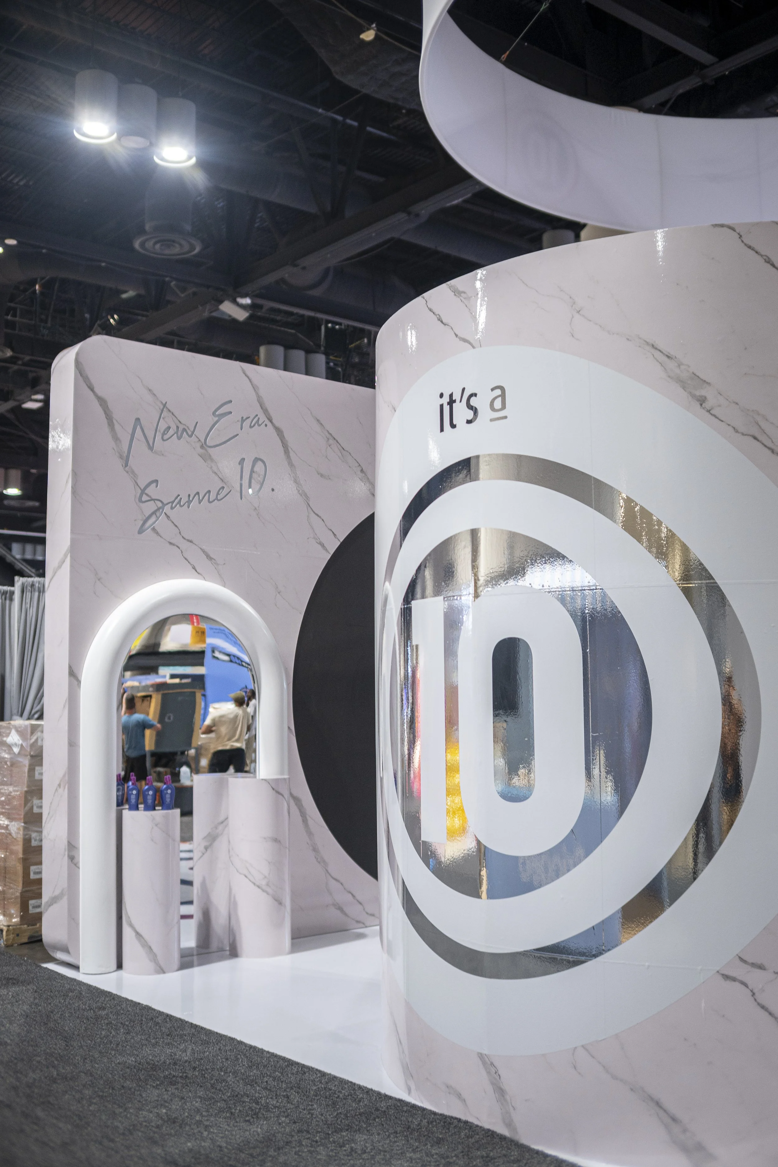 Close-up of a display booth with white marble pattern panels, featuring a large logo with a stylized 'a' inside a circle, and a mirror reflecting people and products at an exhibition.