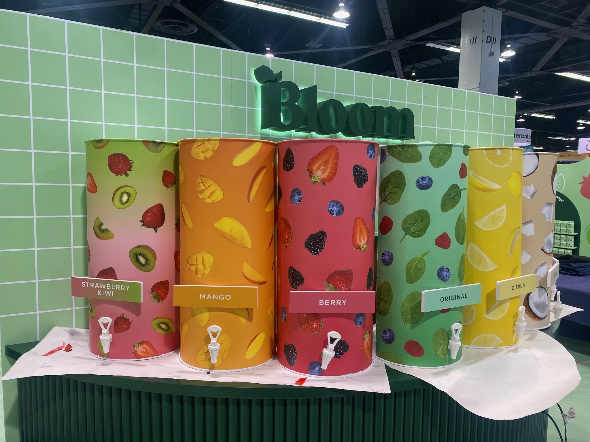 Display of colorful drink dispensers with fruit labels such as strawberry kiwi, mango, berry, and citrus at a store or exhibition.