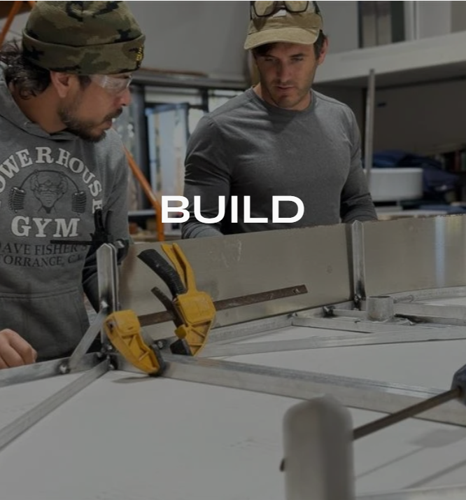Two men in a workshop working on a project with clamps and metal parts, one wearing a camouflage hat and the other a beige cap, with the word BUILD over the image.