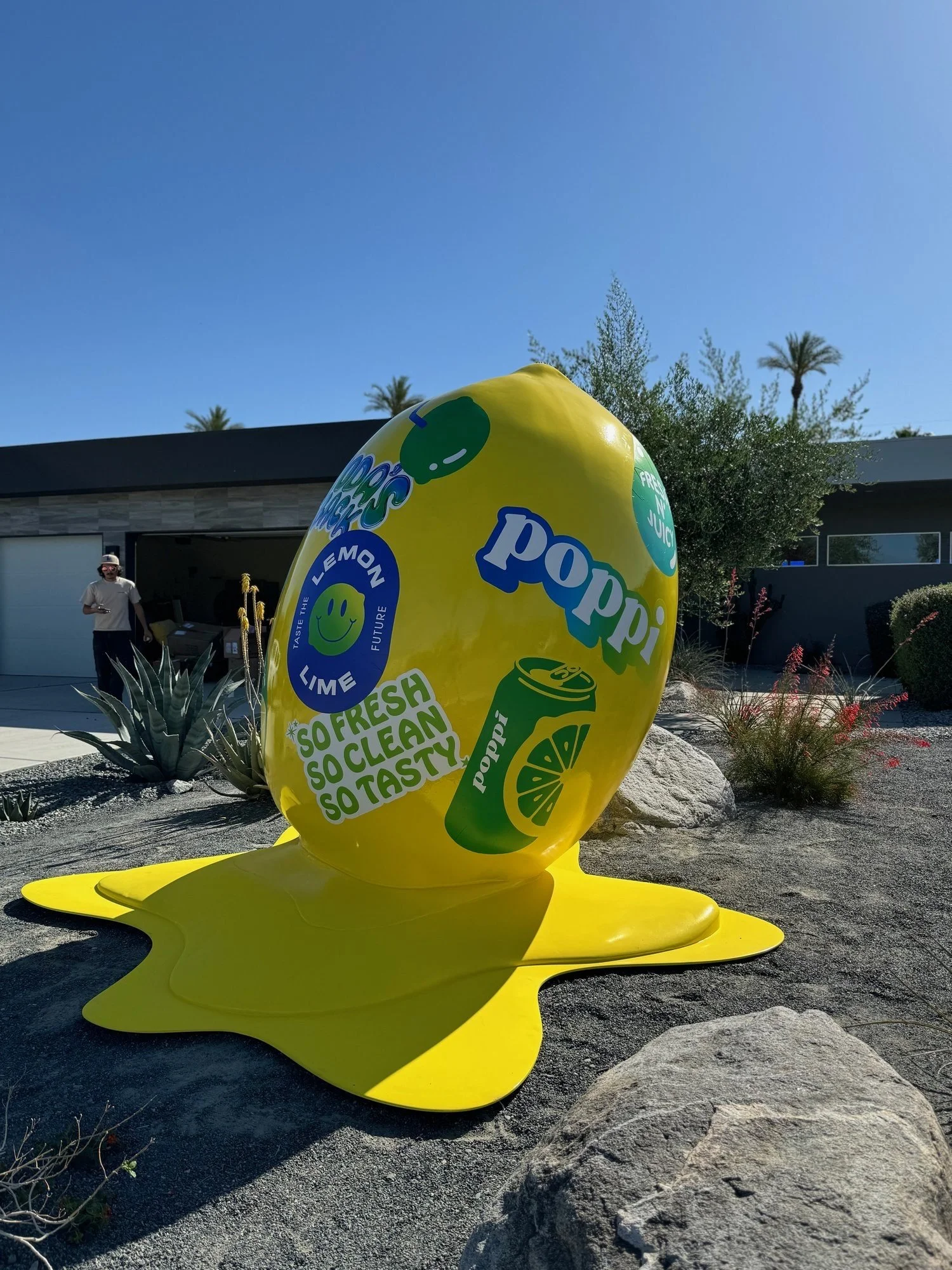 Large yellow inflatable lemon with green accents and playful text, laid on the ground in an outdoor desert-style landscape with rocks, plants, and trees, and a woman standing in the background.