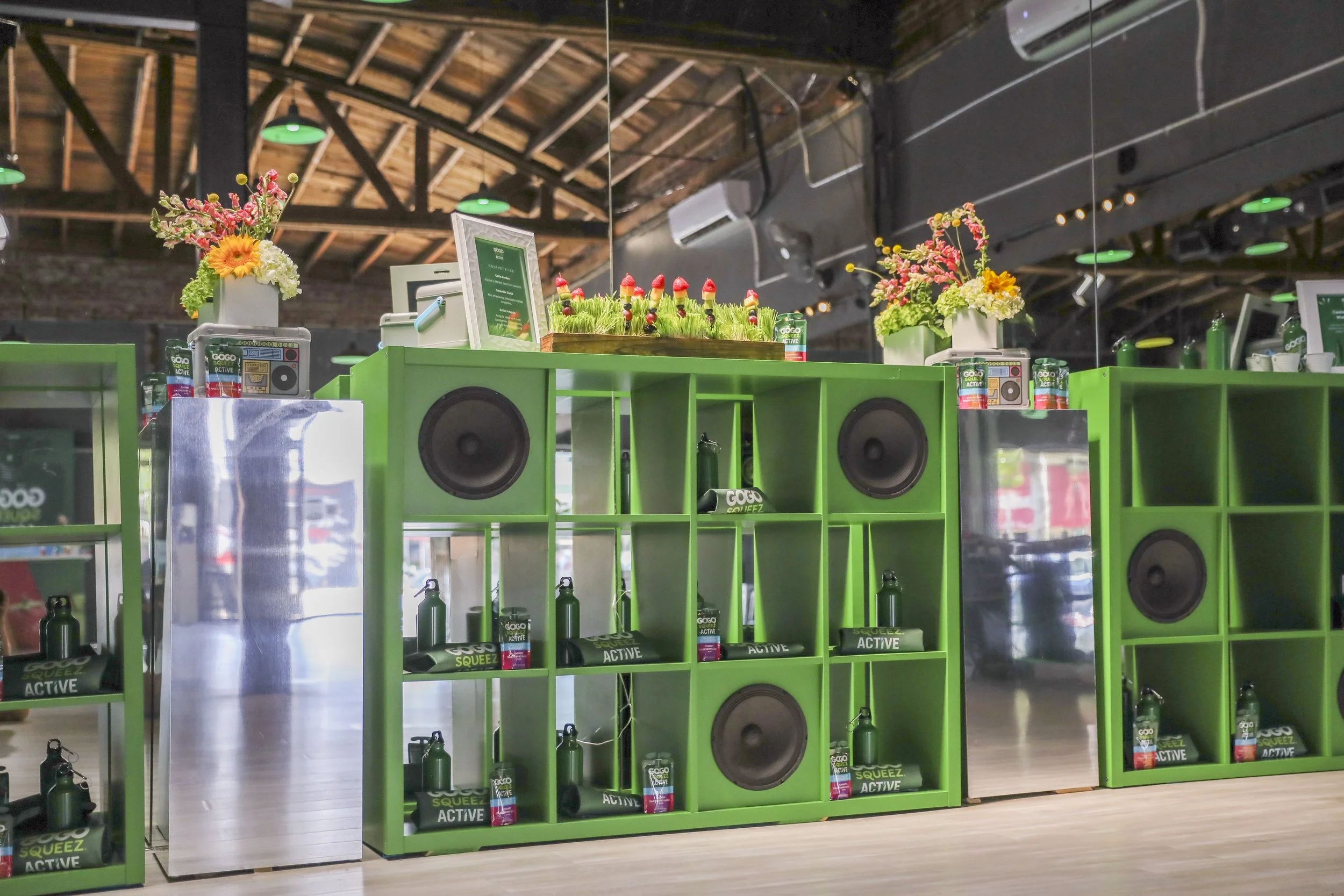 Green display shelves with bottles of GOGO SQUEEZ ACTIVE and other products, decorated with flower arrangements, in a store with exposed wooden ceiling beams and air conditioning units.