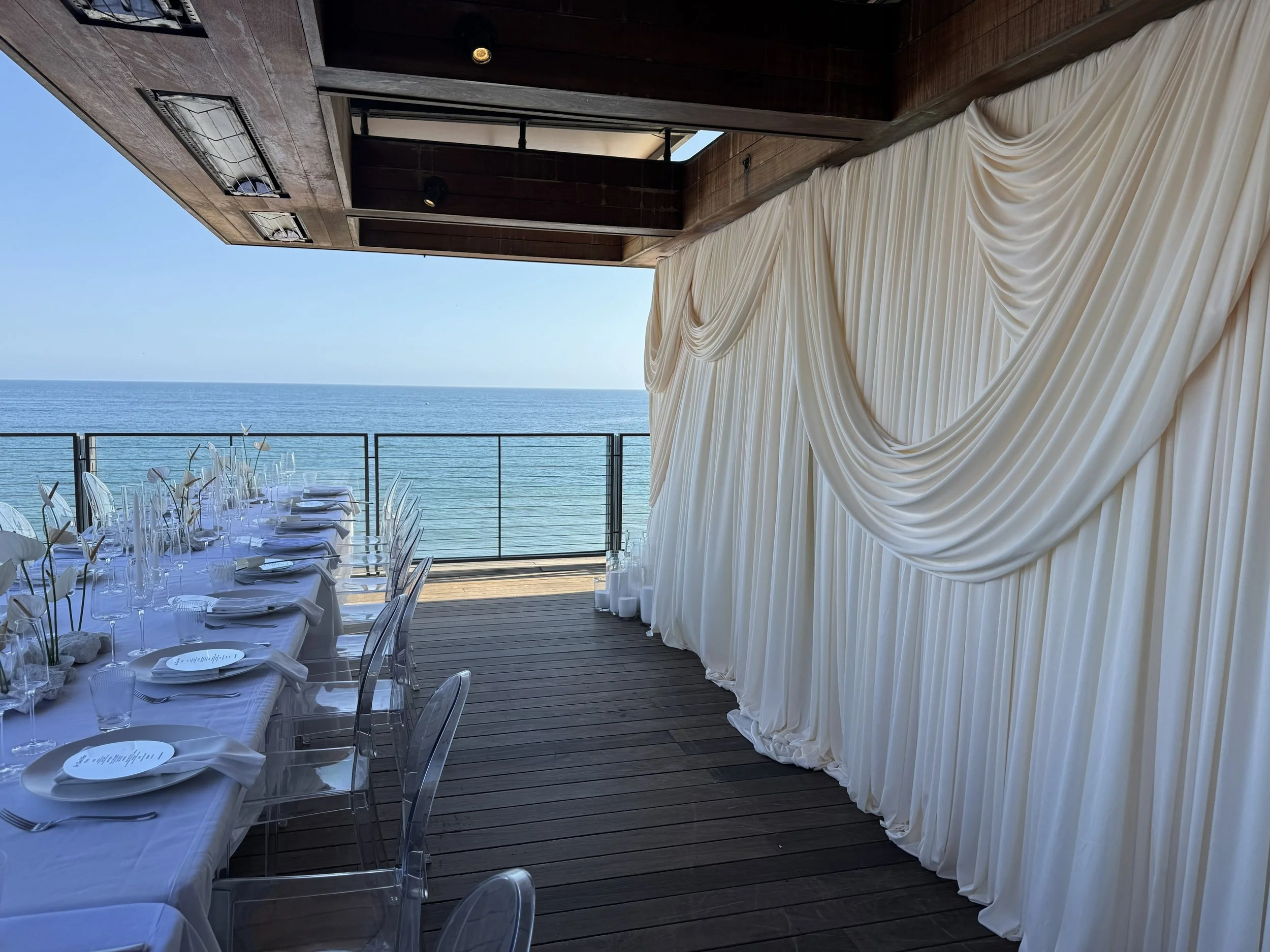 A seaside wedding reception setup on a terrace with a view of the ocean, featuring long tables with white tablecloths, clear chairs, and elegant place settings. There's a large white draped fabric backdrop on the right and a wooden ceiling, with the 