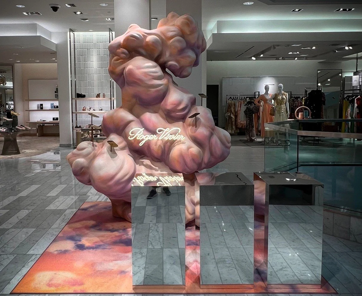 Sculpture of a cloud-like form with pastel pink, purple, and orange hues, with the signature 'Roger Thore' on it, situated in a retail store with mannequins and clothing racks in the background.